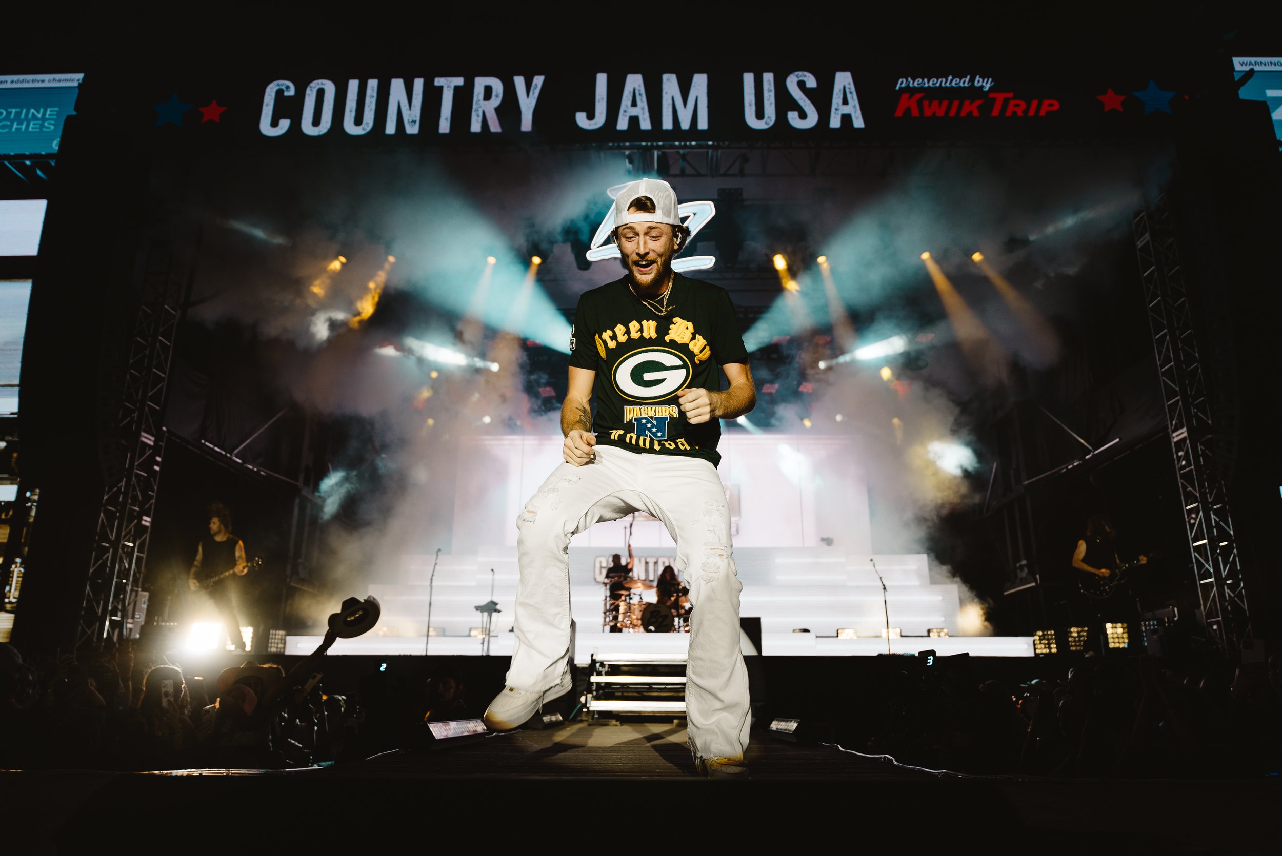 A male performer is dancing on stage at Country Jam USA, wearing a Black Green Bay Packers t-shirt, white pants, and a white backwards cap. The stage has bright lights and smoke effects, with band members playing guitars in the background.