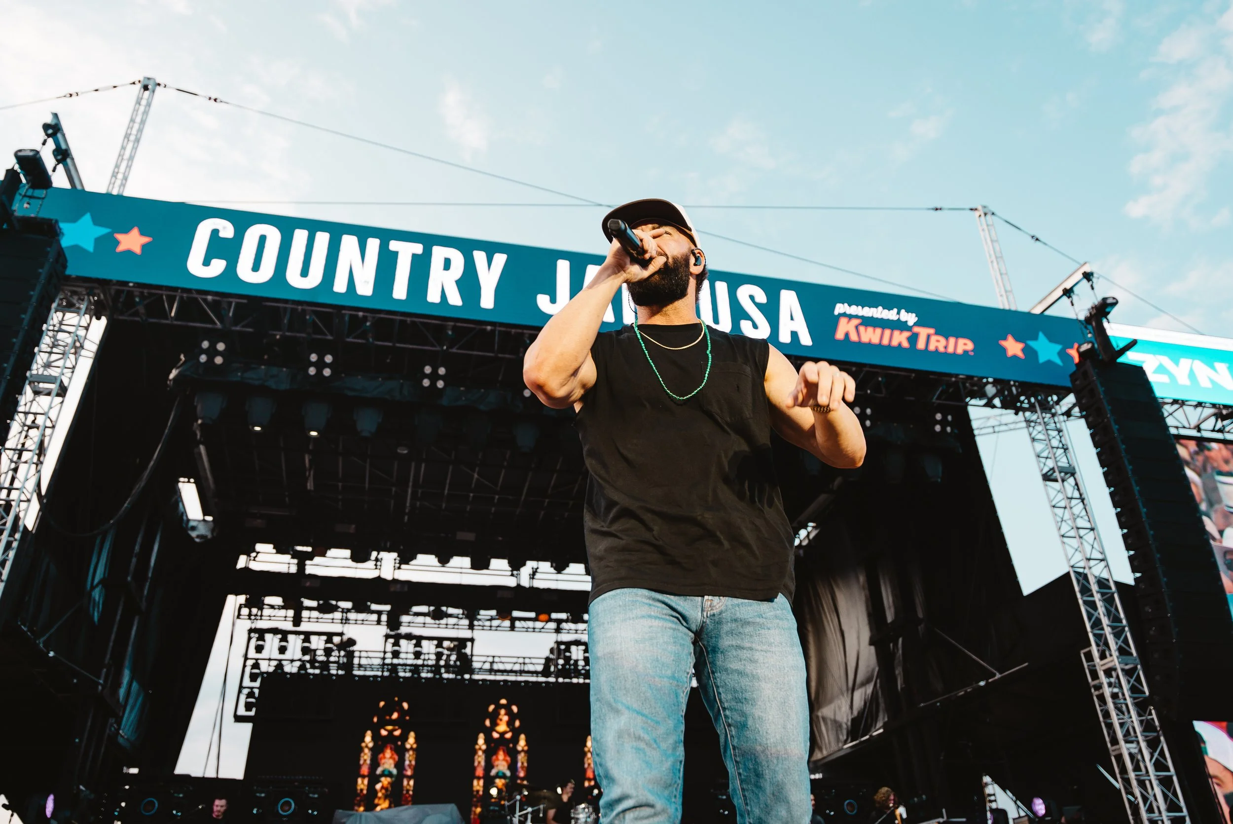 A man with a beard, wearing a black sleeveless shirt, jeans, a baseball cap, and a chain necklace, singing into a microphone on an outdoor stage during a country music festival.