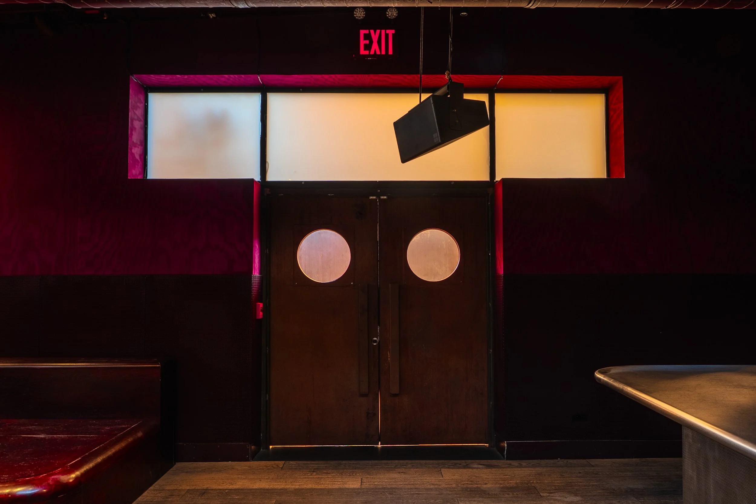 Double wooden door with circular windows in a dimly lit bar or club with black walls, red accents, and a speaker hanging from the ceiling.