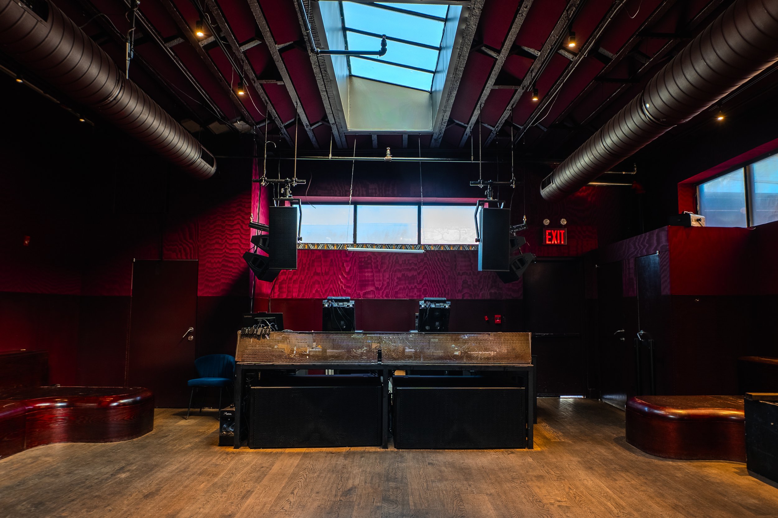 An empty nightclub or concert venue with DJ equipment, large speakers, red walls, wood flooring, and a skylight ceiling.