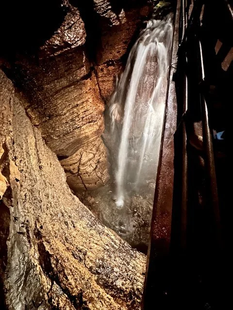 Niagara Cave's underground waterfall plunges into a rocky basin beneath layers of limestone formations. Warm lighting reflects off the water and cave walls, revealing the textures and natural features of the subterranean chamber.
