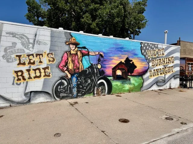 Colorful mural showing John Wayne riding a motorcycle with a covered bridge in the background, featuring the phrases ‘Let’s Go’ on the left and ‘We’re Burning Daylight’ on the right.