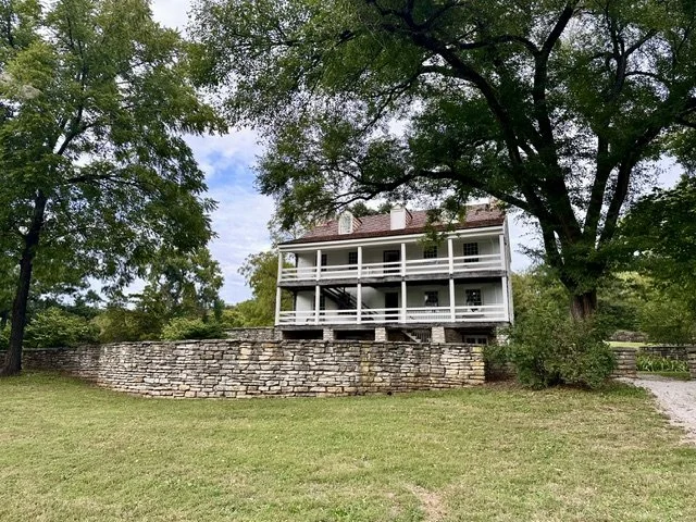 Historic Daniel Boone Home behind a stone retaining wall, surrounded by leafy trees under a blue sky.
