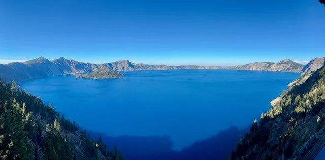 Electric vehicle road trip stop overlooking Crater Lake in Crater Lake National Park, Oregon, with deep blue water surrounded by forested cliffs and clear skies.