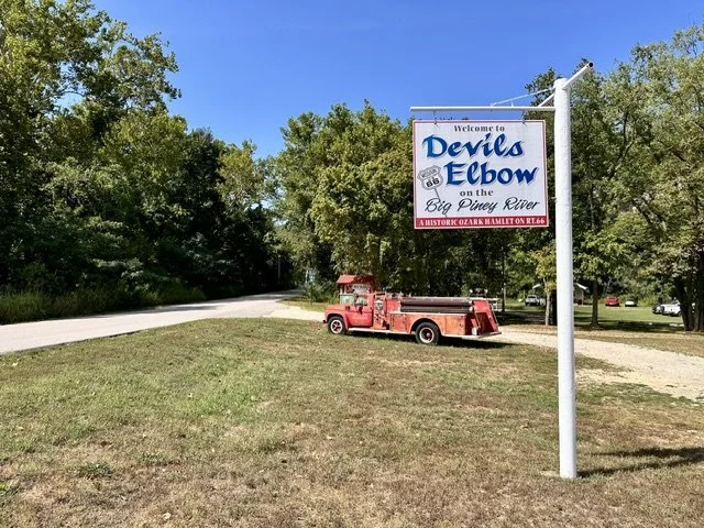 Welcome to Devil's Elbow sign beside historic Route 66 near the Big Piney River in Missouri, with an antique red fire engine parked in a grassy lot under a clear blue sky.
