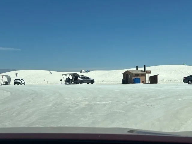 A parking area partially buried in white sand is located within White Sands National Park. Rows of simple picnic shelters line the edge of the lot near the base of the sand dunes, with rolling white dunes rising behind them under a clear, bright sky.