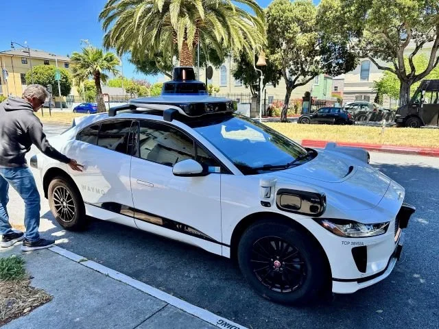 Man opening the rear door of a Waymo autonomous vehicle parked on a street in San Francisco, showcasing self-driving car technology in urban environment.