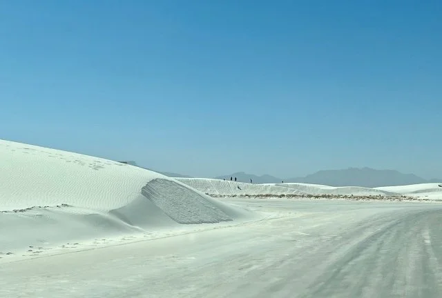 A paved roadway partially covered in white sand stretches through White Sands National Park. Soft, wave-like sand dunes line both sides of the road, and a mountain range can be seen in the distance under a bright, open sky.