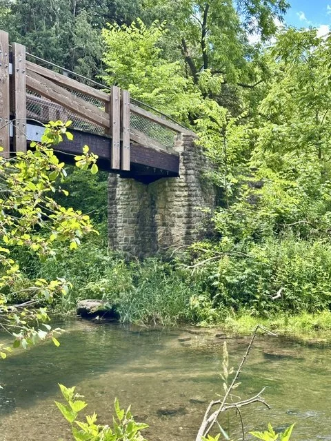 Civilian Conservation Corps-built stone bridge crossing the Whitewater River in Whitewater State Park, surrounded by full green foliage on a sunny day with a blue sky and scattered clouds.