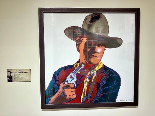 1985 Andy Warhol pop‑art painting of John Wayne wearing a cowboy hat and holding a six‑shot revolver pistol, featuring bold colors and Warhol’s signature graphic style.