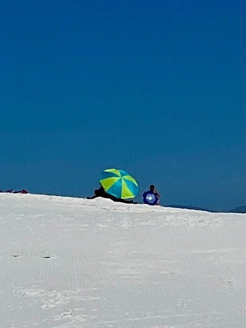 A white gypsum sand dune rises in White Sands National Park, where a small group of people sit together under a blue and green beach umbrella for shade. The smooth, sunlit dune contrasts with the colorful umbrella against a clear sky.