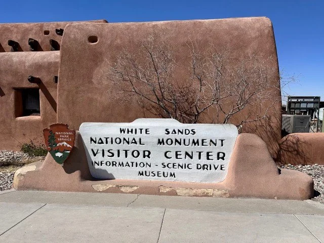 A large entrance sign reading "White Sands National Monument Visitor Center" is positioned in front of a low, adobe-style ranger station and gift shop building. The scene shows a typical bright southern New Mexico sky.