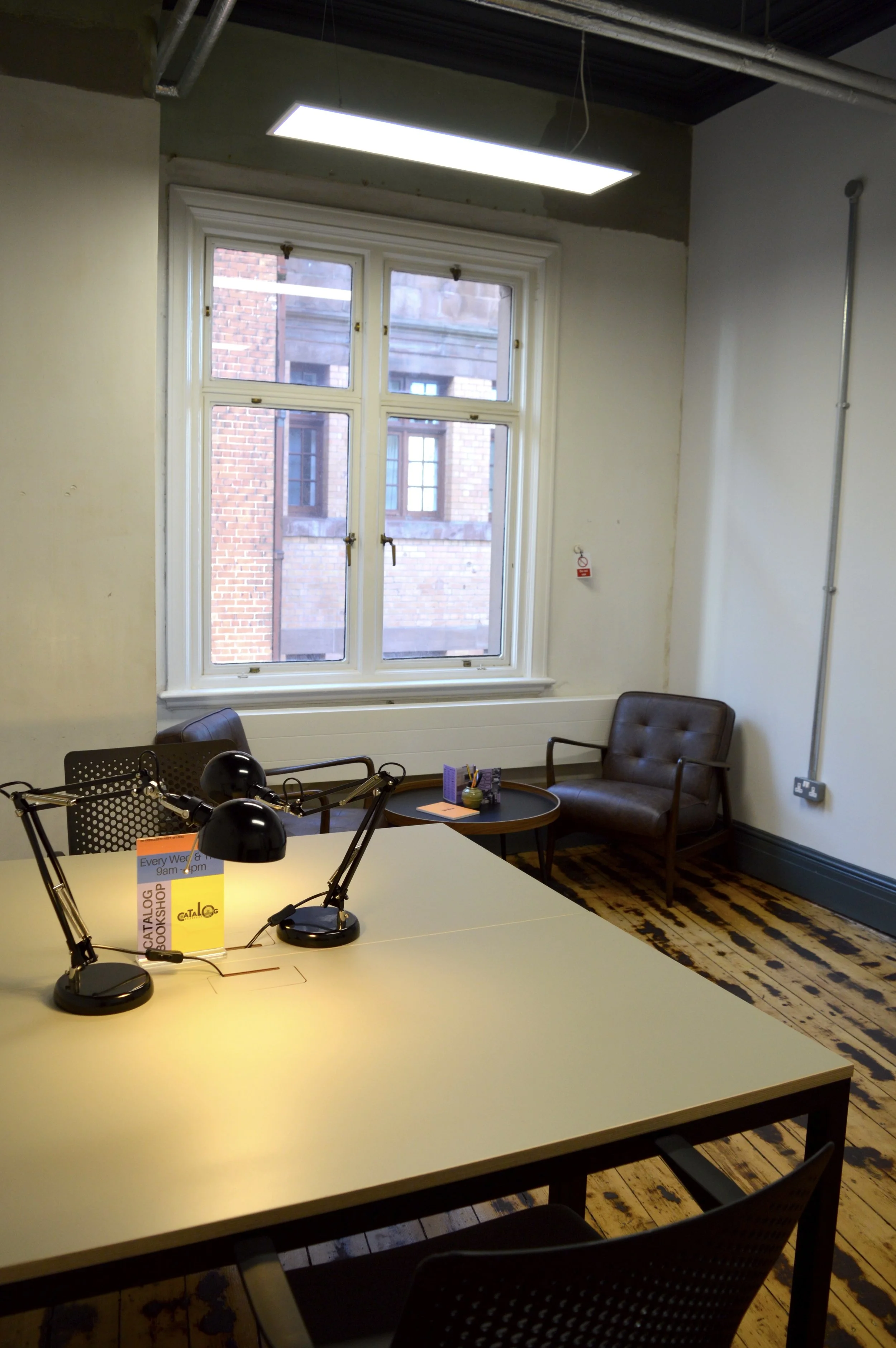 A private studio in SEESAW featuring a desk, a comfy corner with leather chairs by the window with a view of Manchester's red brick architecture outside.