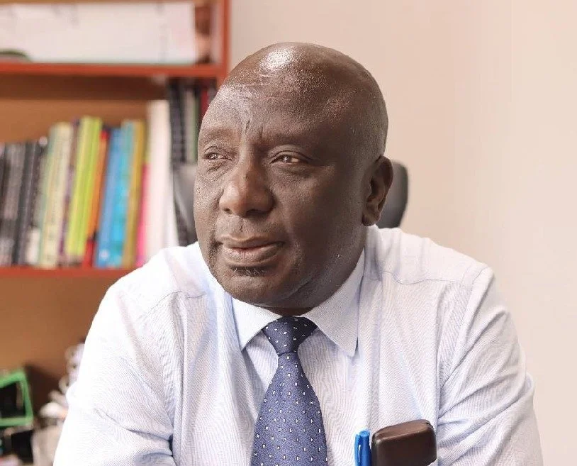 A middle-aged man with dark skin and a bald head, wearing a light blue collared shirt and a navy tie with small white dots, sitting in an office with a bookshelf in the background.
