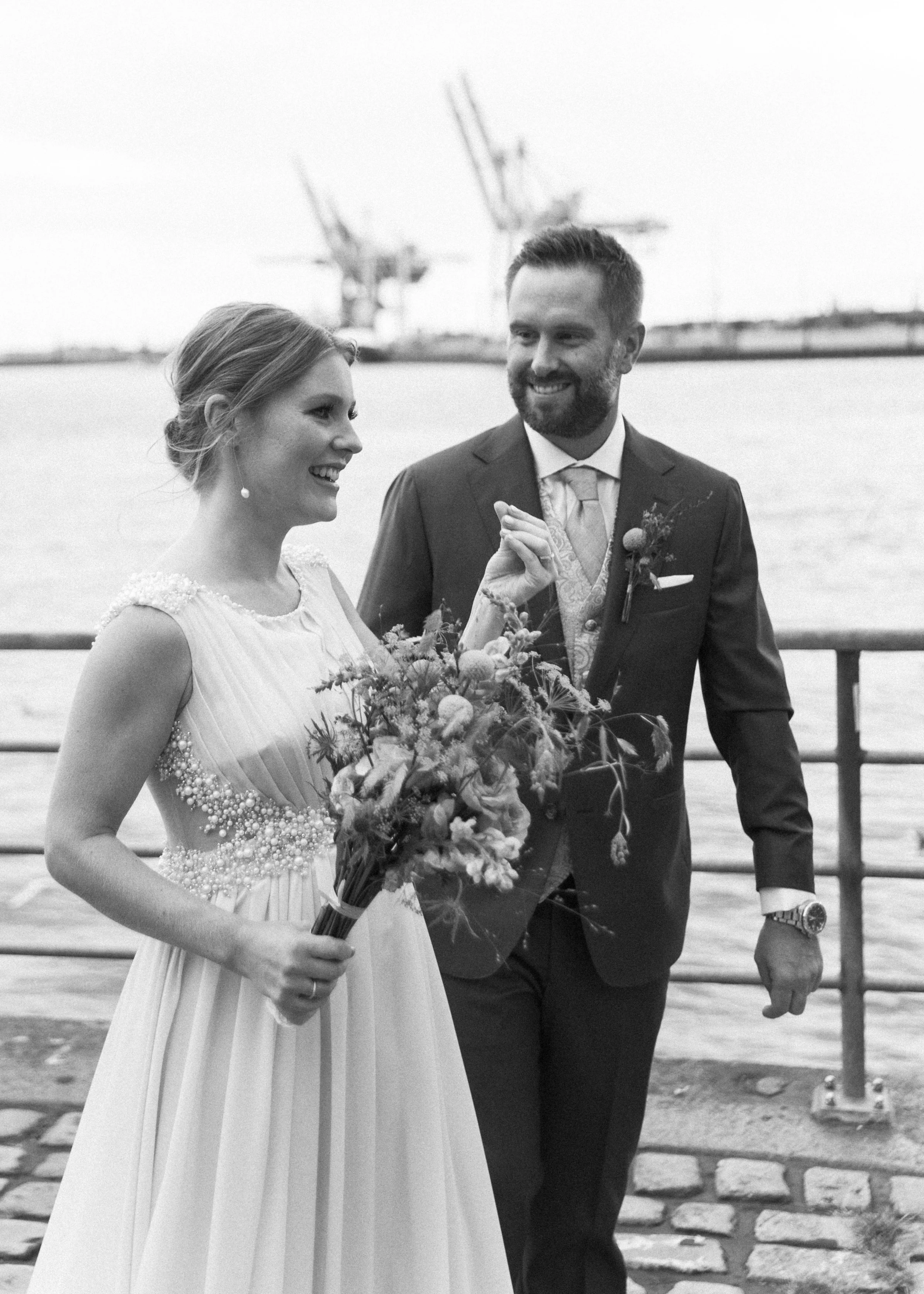 Braut und Bräutigam bei ihrer Hochzeit am Wasser, lachend, mit Hochzeitsstrauß, im Hintergrund Hafencranes