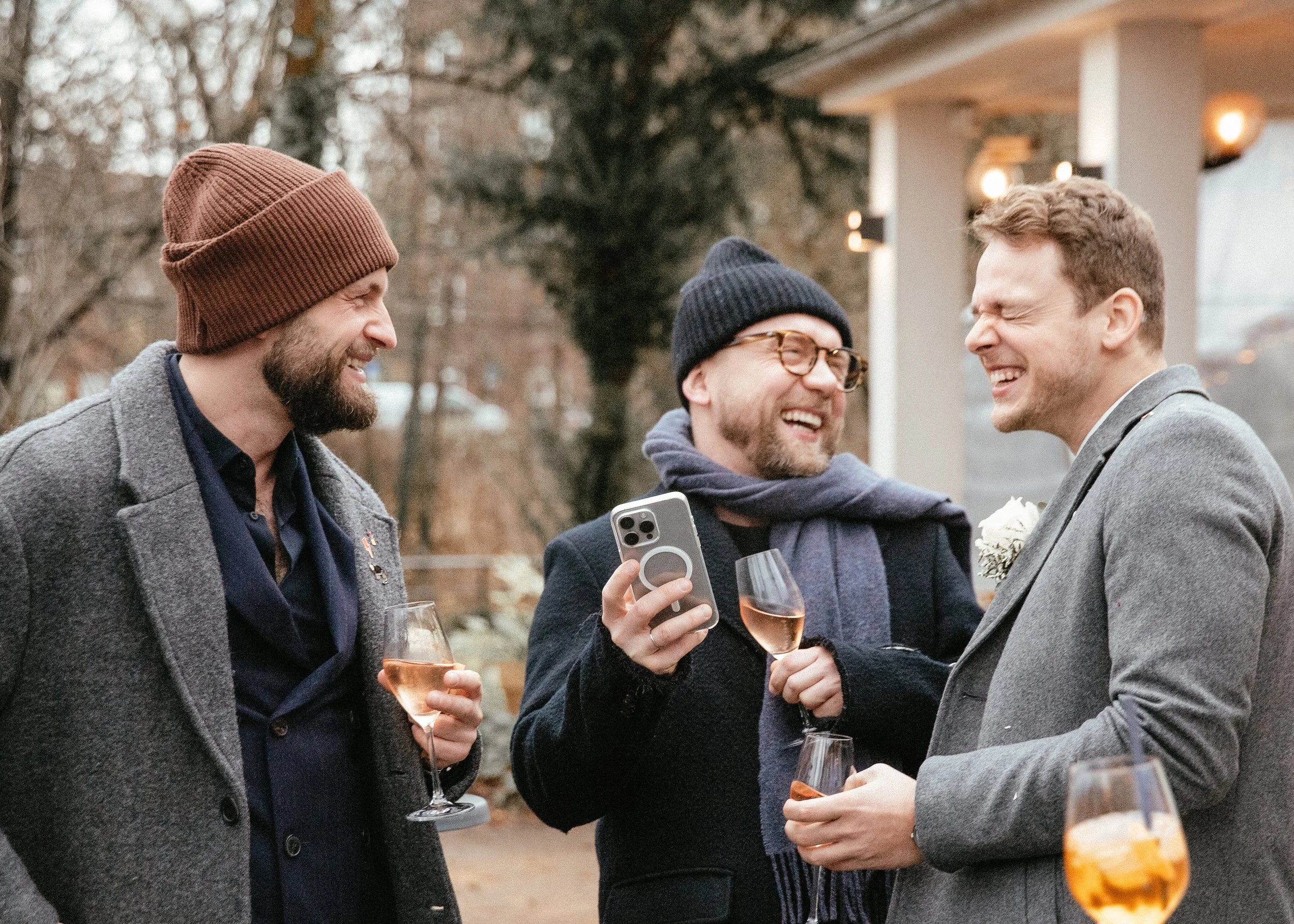 Drei Männer im Freien, die bei einer Feier lachen, mit Weingläsern und einem Handy, die sich scheinbar amüsieren und unterhalten.