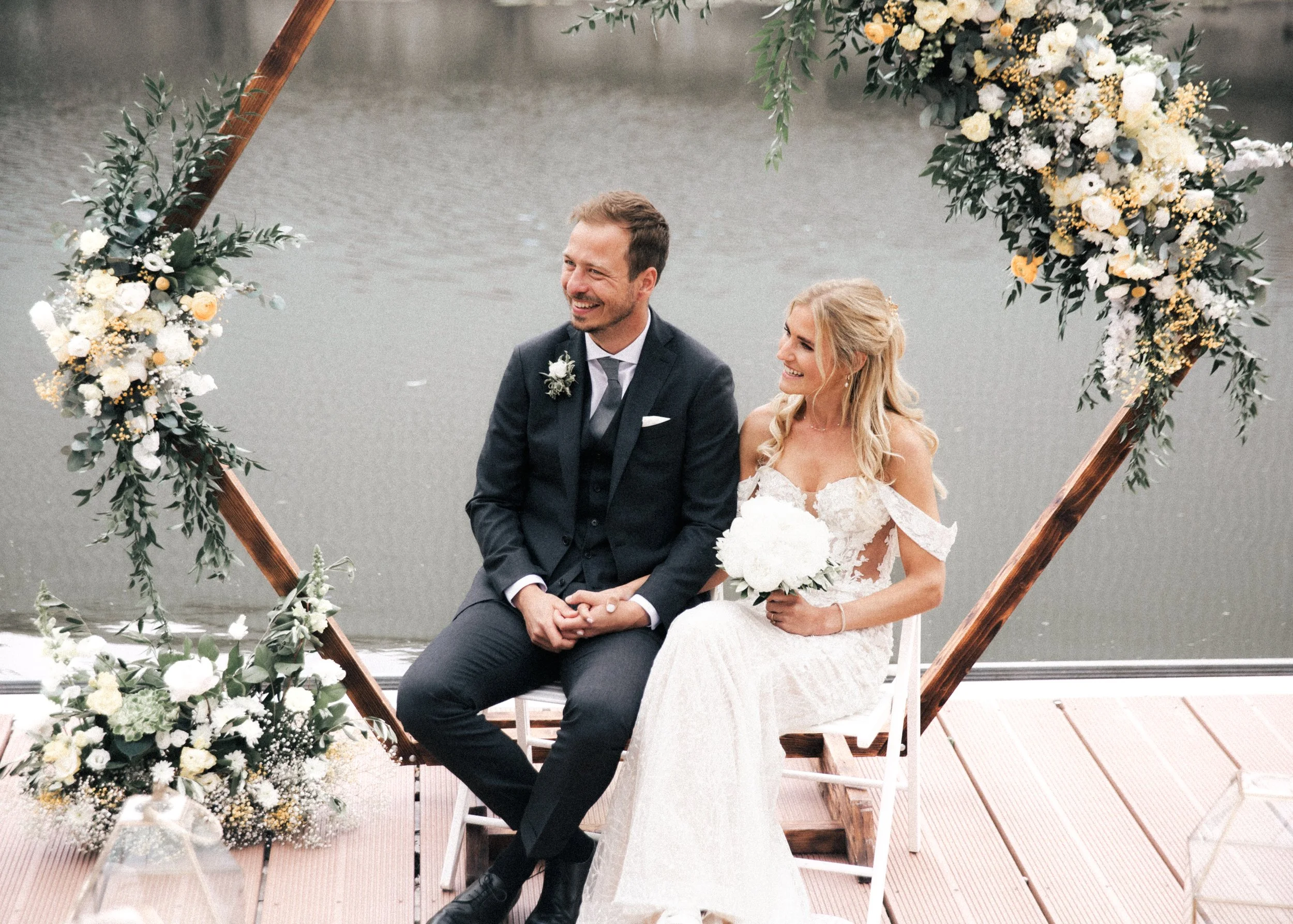 Ein glückliches Brautpaar sitzt an einem Steg an einem Fluss, umgeben von Blumen, bei einer Hochzeit.