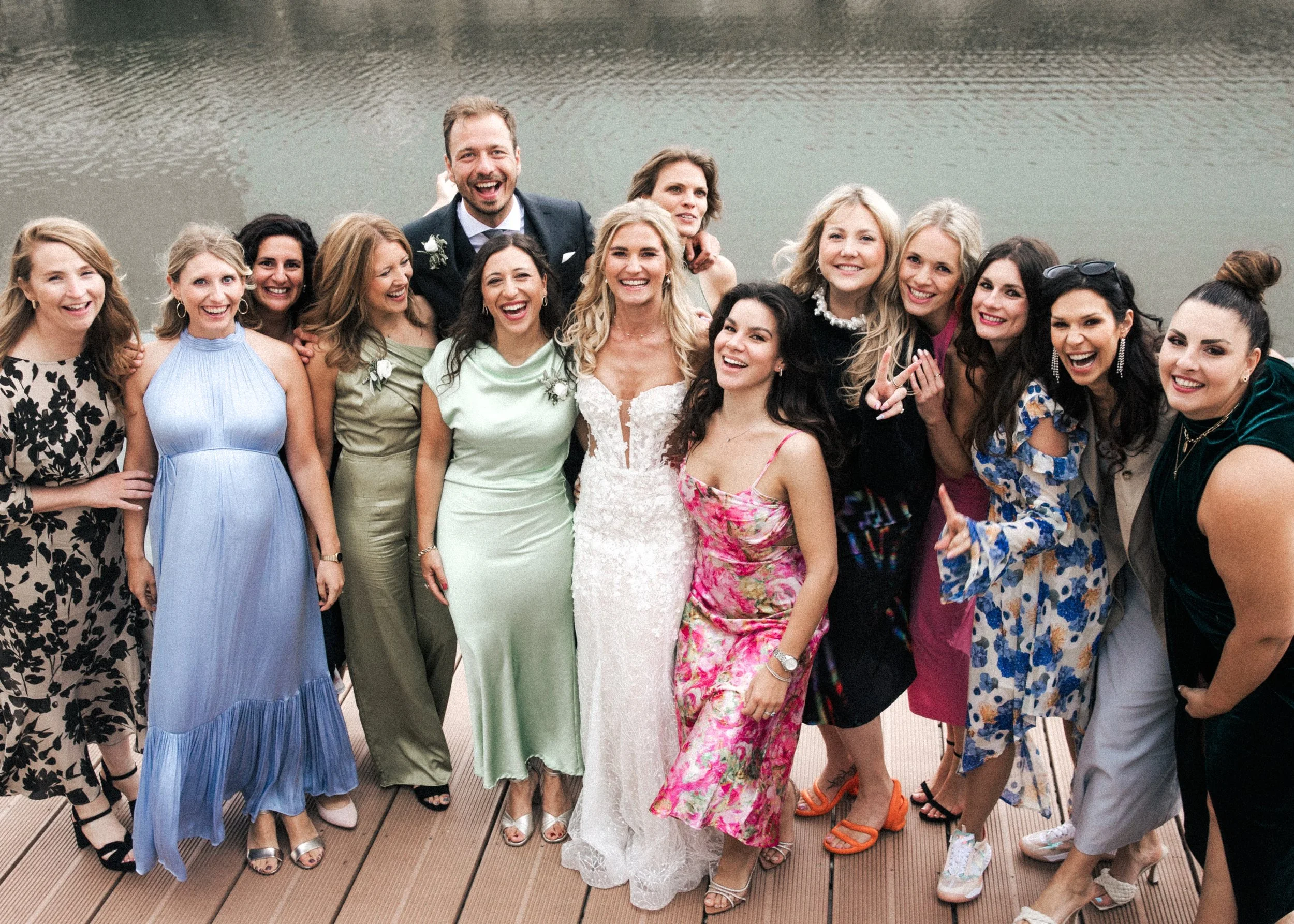 Mehrere Frauen und ein Mann in festlicher Kleidung stehen am Wasser auf einer Brücke, sie lachen und posieren für ein Gruppenfoto bei einer Hochzeit.
