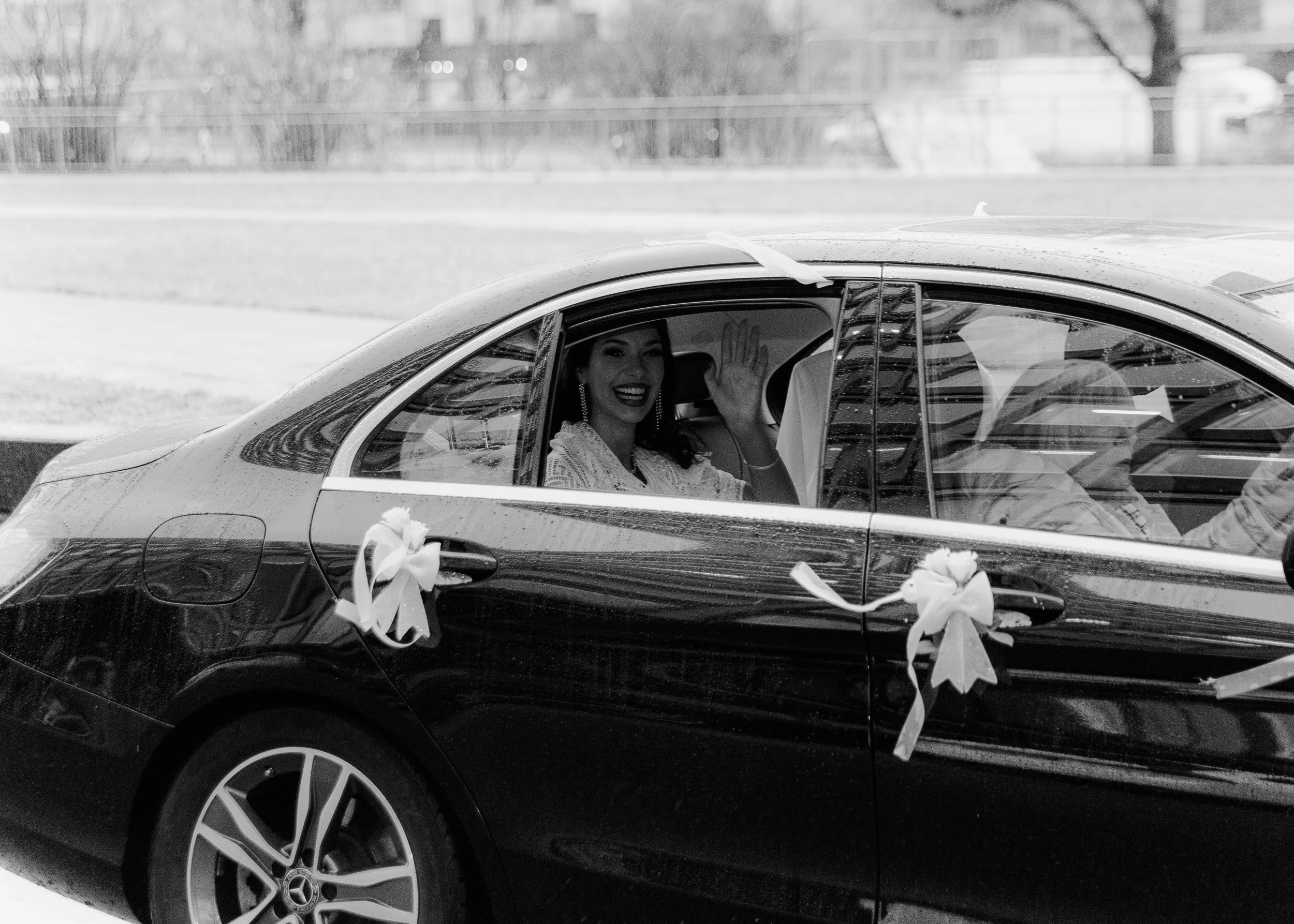 Eine lächelnde Frau sitzt in einem schwarzen Auto, winkt und trägt elegante Kleidung. Das Auto ist mit Schleifen und Dekorationen für eine festliche Gelegenheit geschmückt.