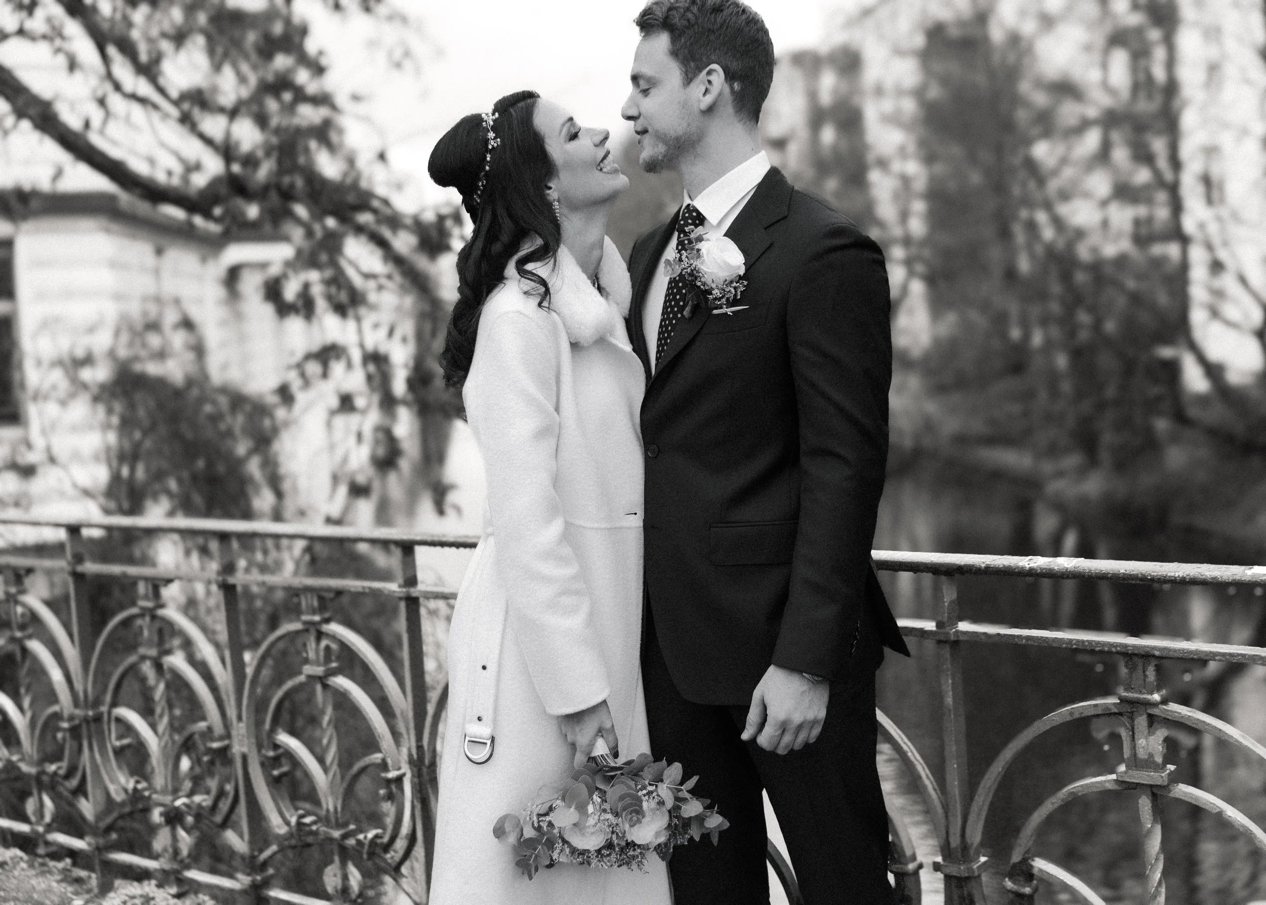 Ein Brautpaar steht sich an einem Brückenrand gegenüber, lächelnd, im Hintergrund Bäume und ein Fluss in einem Park, schwarz-weiß Fotografie.