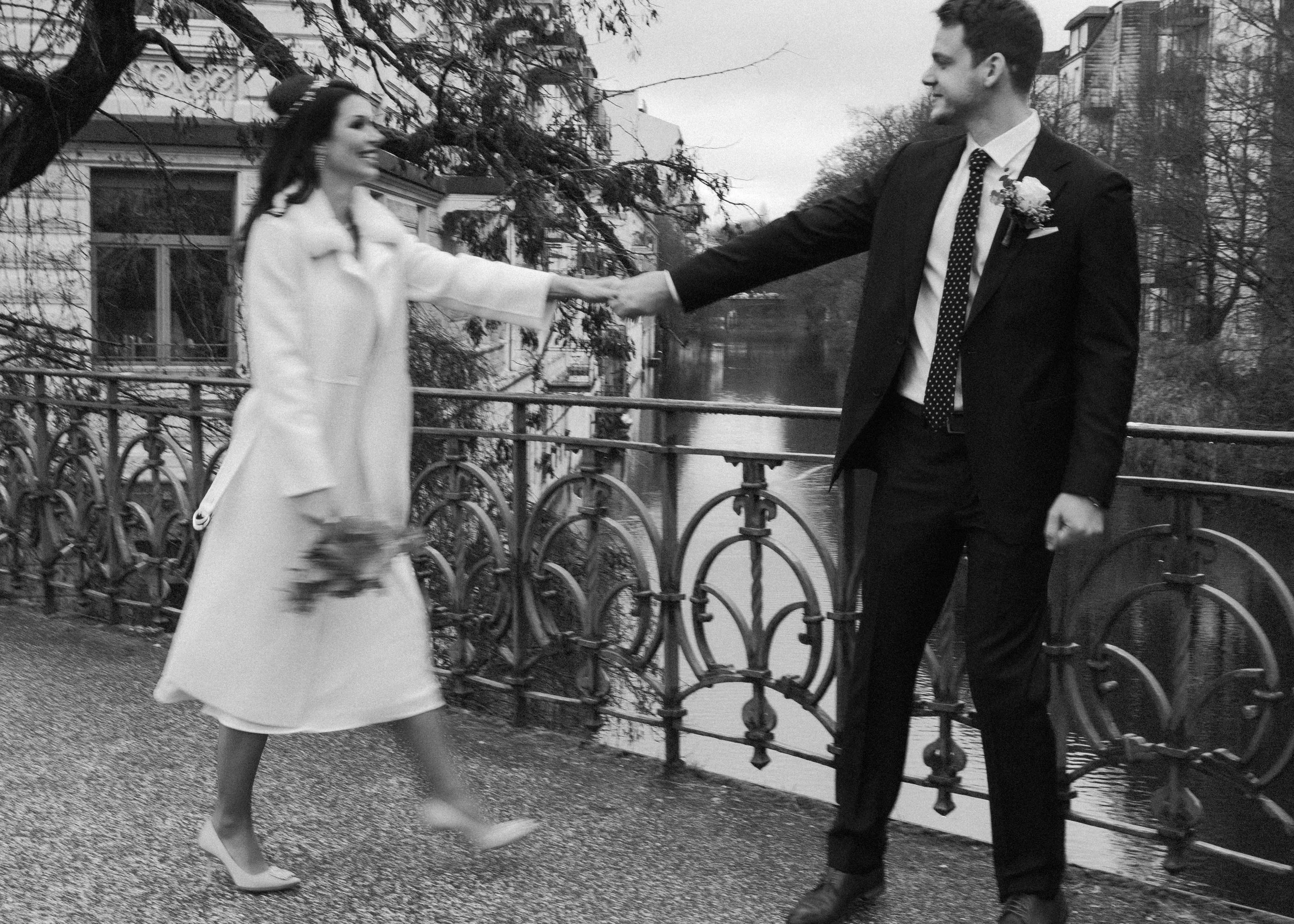 Ein Mann in Anzug und eine Frau in weißem Mantel, die eine Hand hält, stehen auf einer Brücke neben einem Fluss, im Hintergrund Bäume und Gebäude, schwarz-weiß, romantische Stimmung.