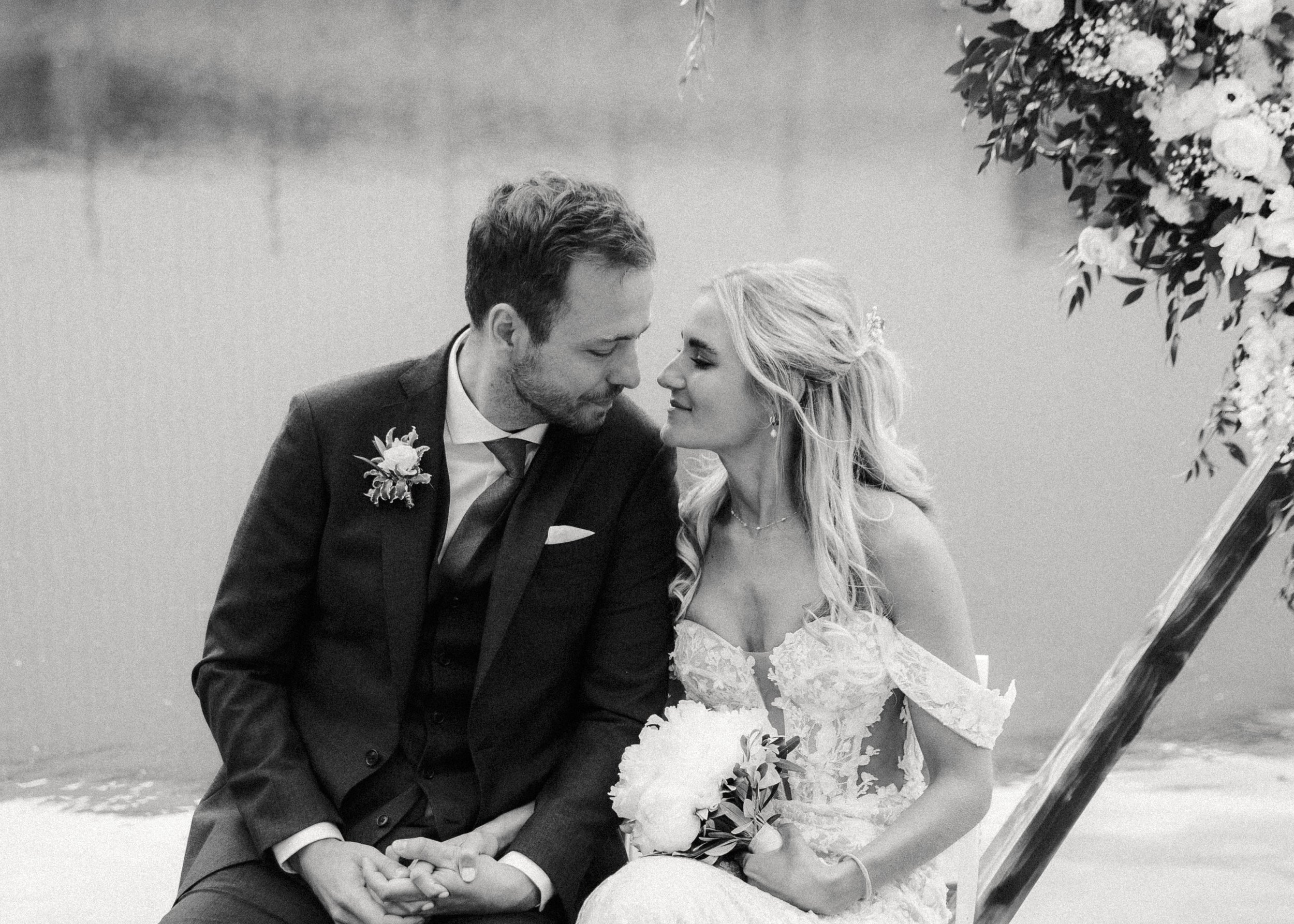 Ein Brautpaar sitzt am Wasser, die Braut hält einen Blumenstrauß, sie blicken sich liebevoll an.