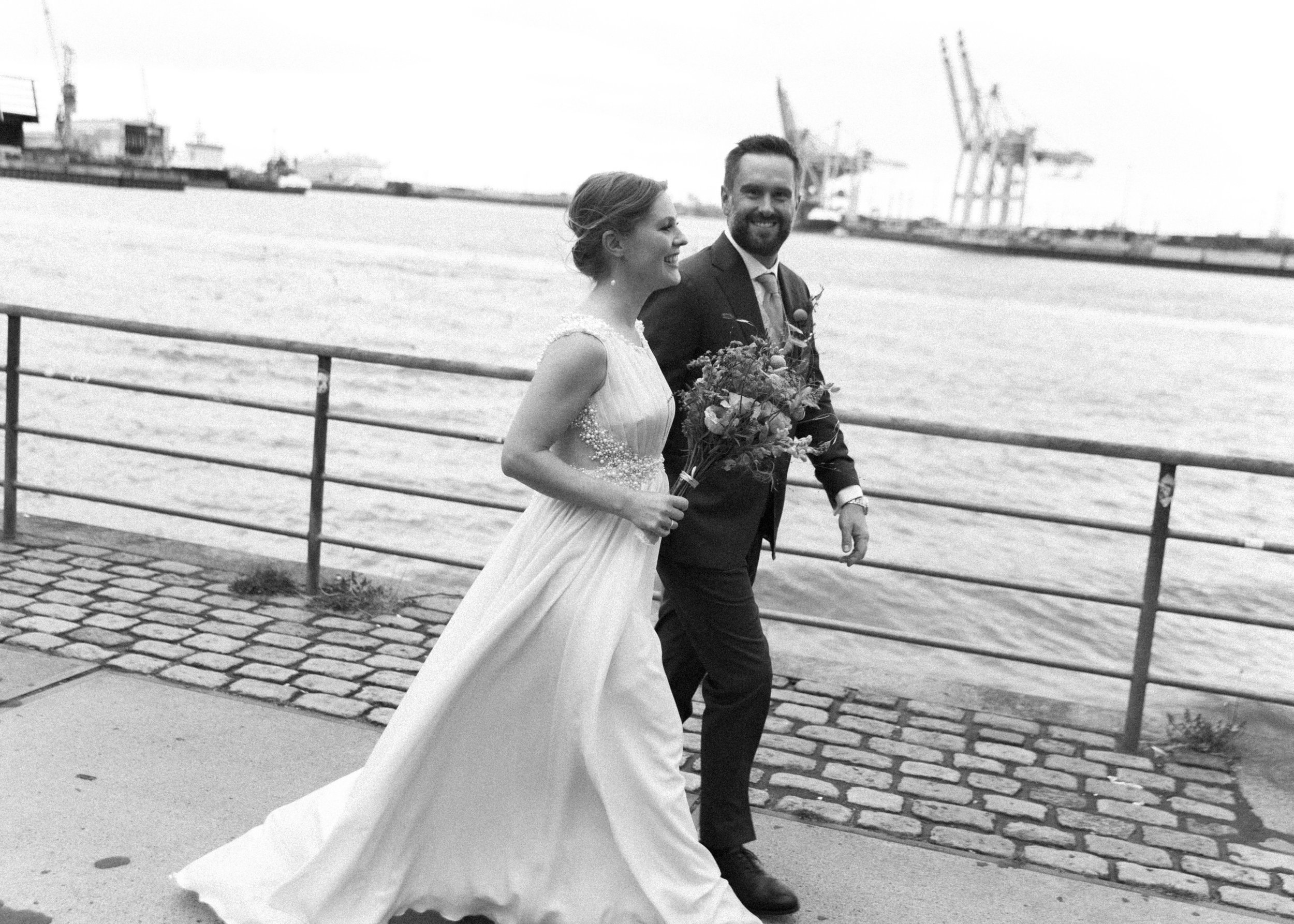 Ein Brautpaar, das am Wasser entlangspaziert, die Braut in einem weißen Kleid mit Blumen und der Bräutigam in einem Anzug, lachend und glücklich.