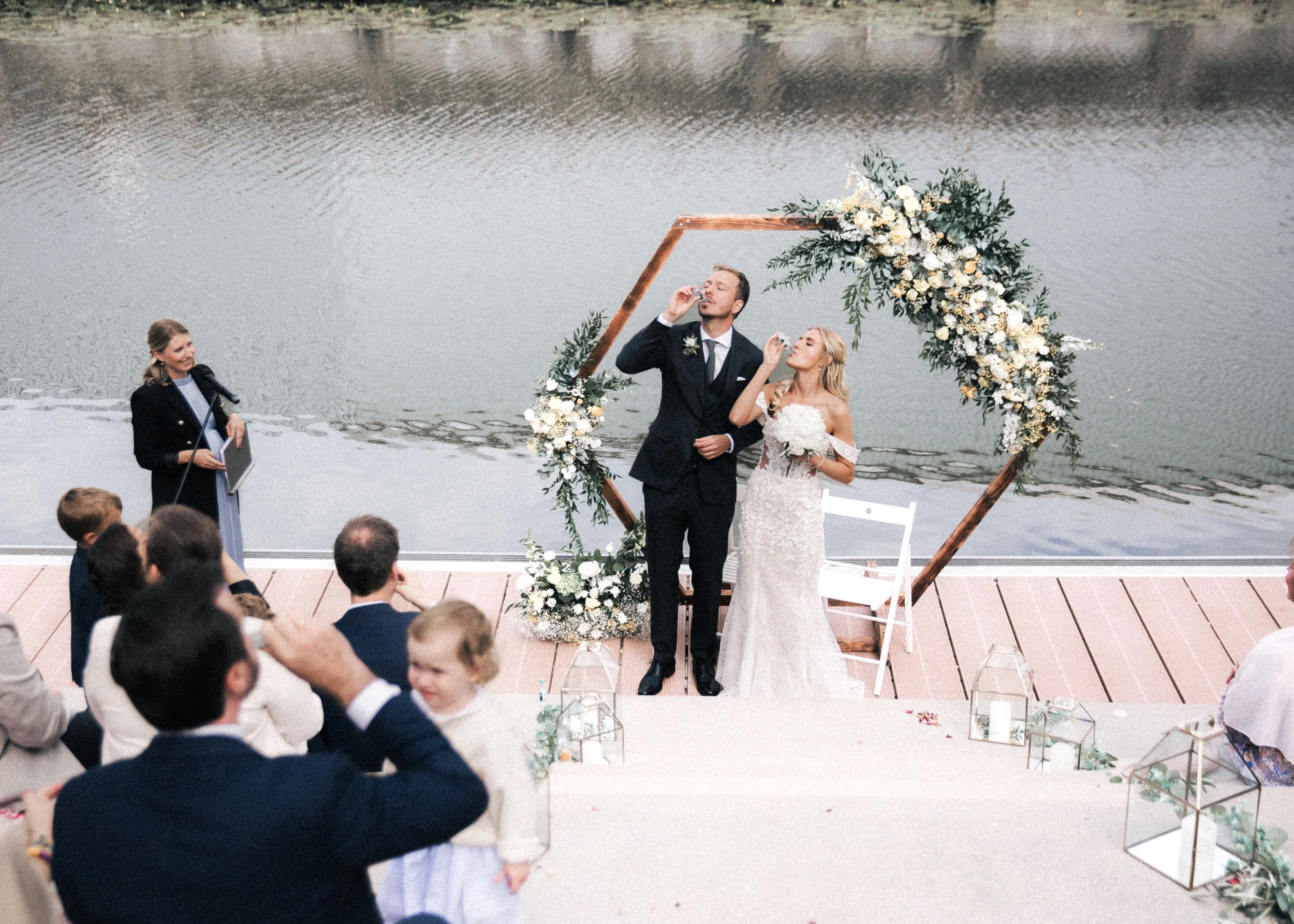 Ein Hochzeitsritual am Wasser mit einigen Gästen, einem Bräutigam und einer Braut vor einem dekorierten hexagonalen Holzrahmen mit Blumen, während eine Hochzeitsrede gehalten wird.