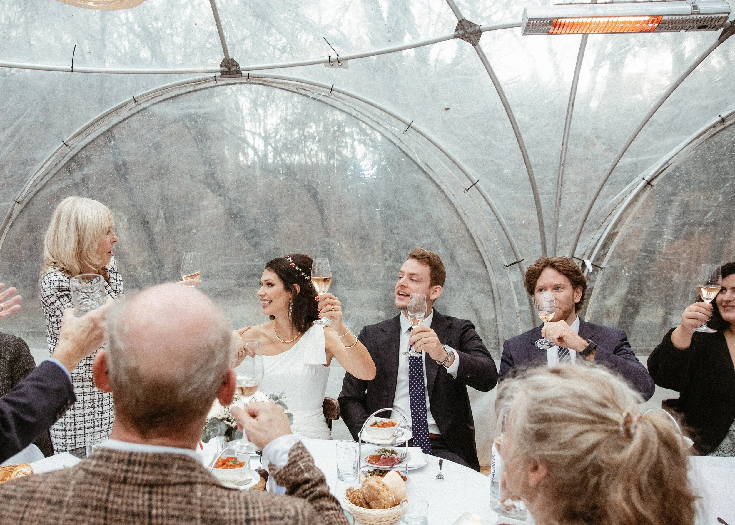 Menschen bei einer festlichen Feier im Zelt, trinken Wein und lachen, festliche Kleidung, weibliche und männliche Gäste, gedeckter Tisch mit Essen und Getränken.