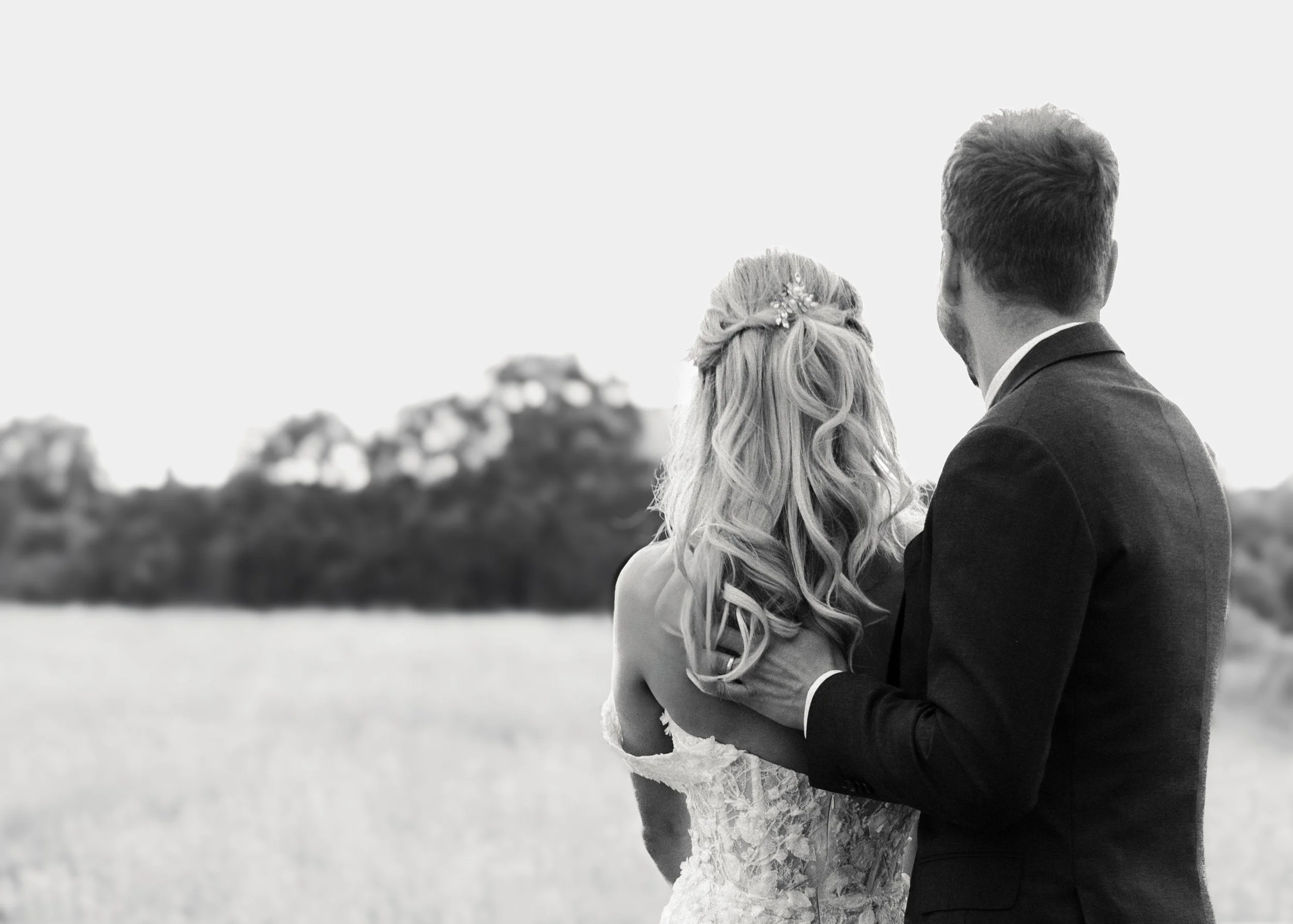 Ein Paar bei einer Hochzeitsfeier im Freien, die Braut trägt ein weißes Kleid mit Spitzen und hat lange, lockige Haare, der Bräutigam trägt einen schwarzen Anzug. Sie stehen in einer Naturumgebung mit Bäumen im Hintergrund.