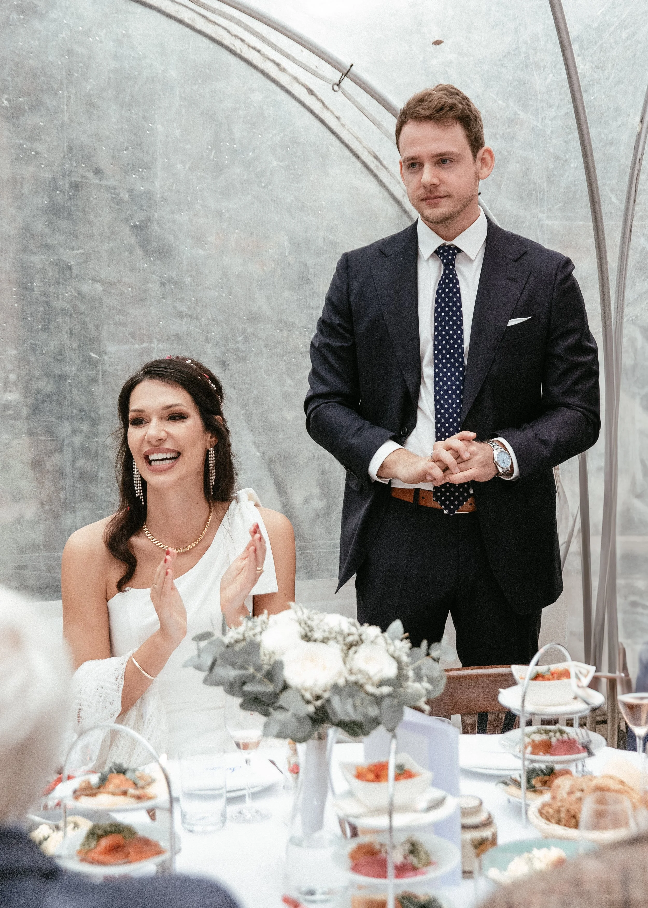 Ein Mann und eine Frau sitzen an einem festlich gedeckten Tisch bei einer Feier. Die Frau ist in einem weißen Kleid und trägt Schmuck, während der Mann einen dunklen Anzug mit Krawatte trägt.