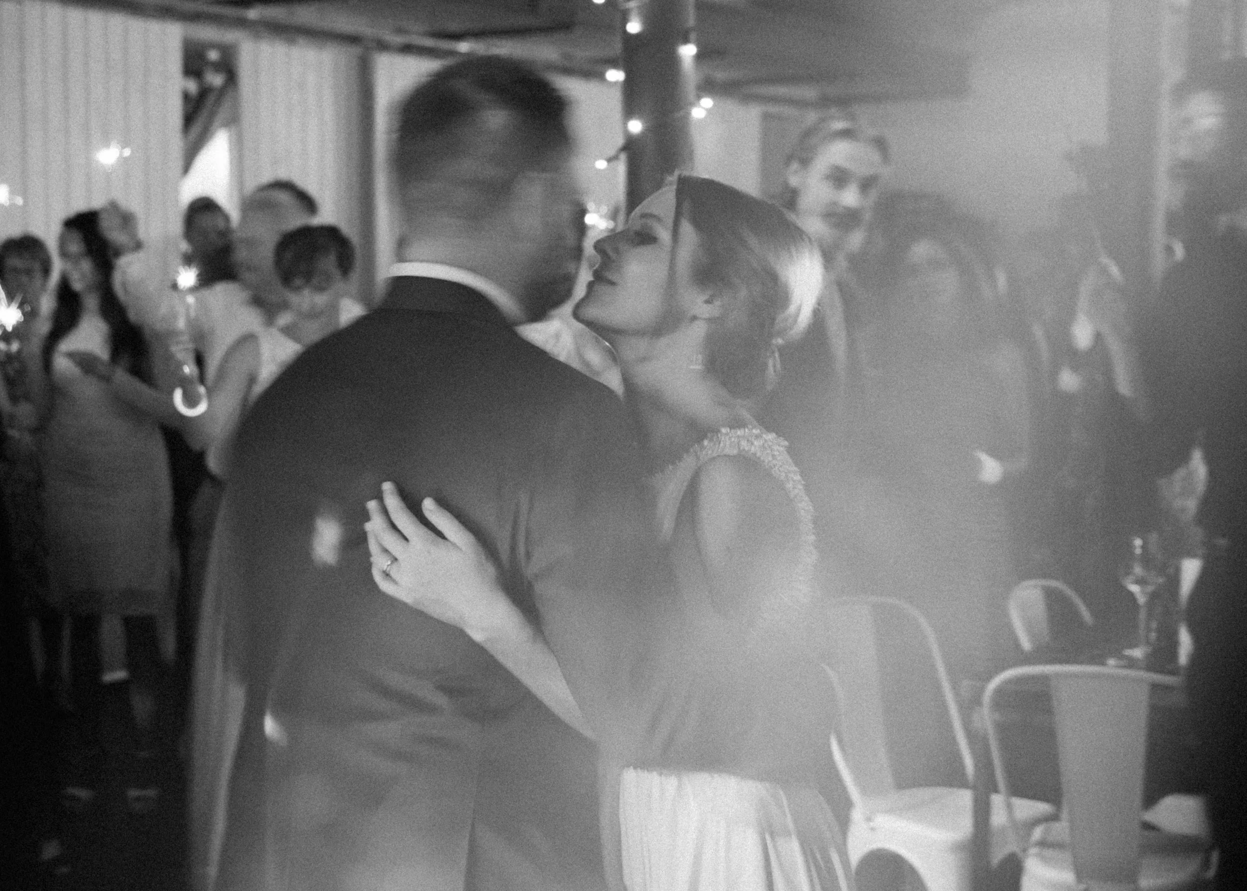Ein Paar tanzt in einem festlichen Raum, umgeben von weiteren tanzenden und stehenden Personen, in Schwarz-Weiß.