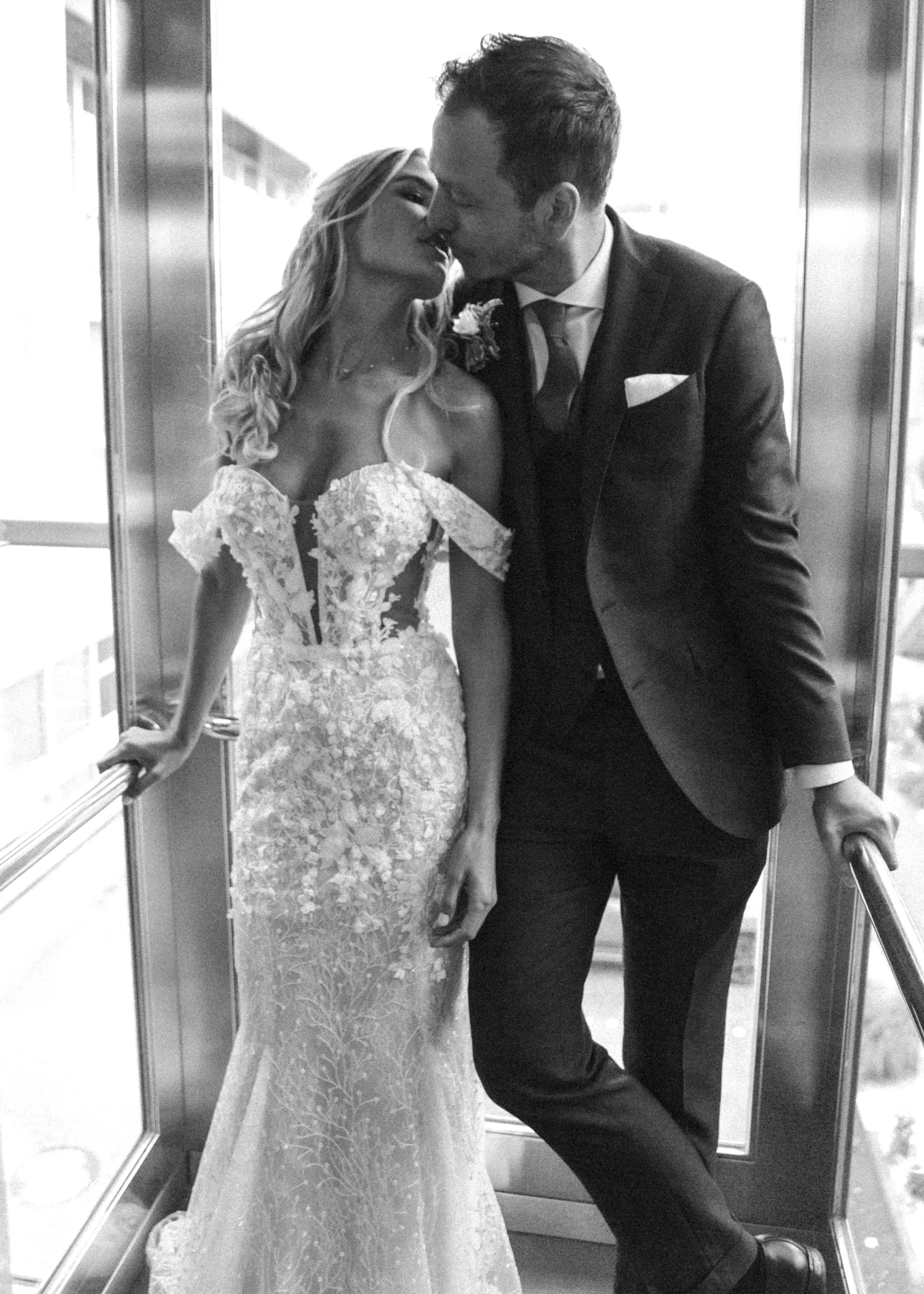 Ein Brautpaar in formeller Kleidung in einem Aufzug, sie küsst sich, er trägt einen dunklen Anzug, sie ein langes, elegantes Hochzeitskleid.