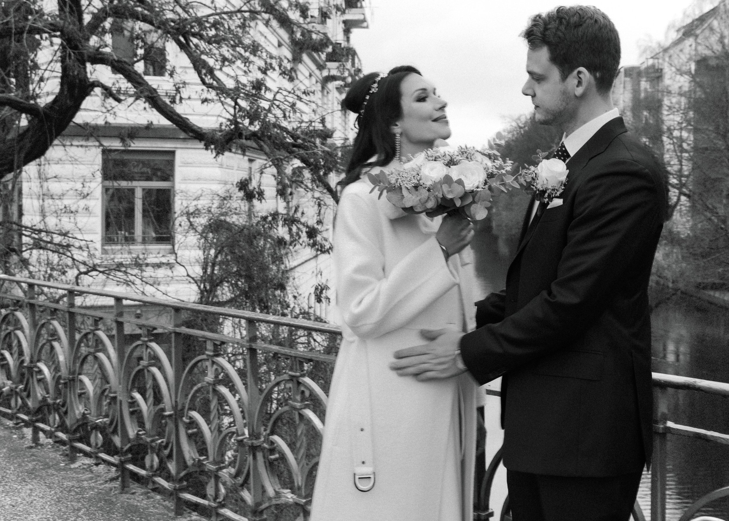 Ein Hochzeitspaar steht am Flussufer, sie lächelt und hält einen Blumenstrauß, er schaut liebevoll auf sie, im Hintergrund Bäume und ein altes Gebäude, schwarz-weiß Foto.