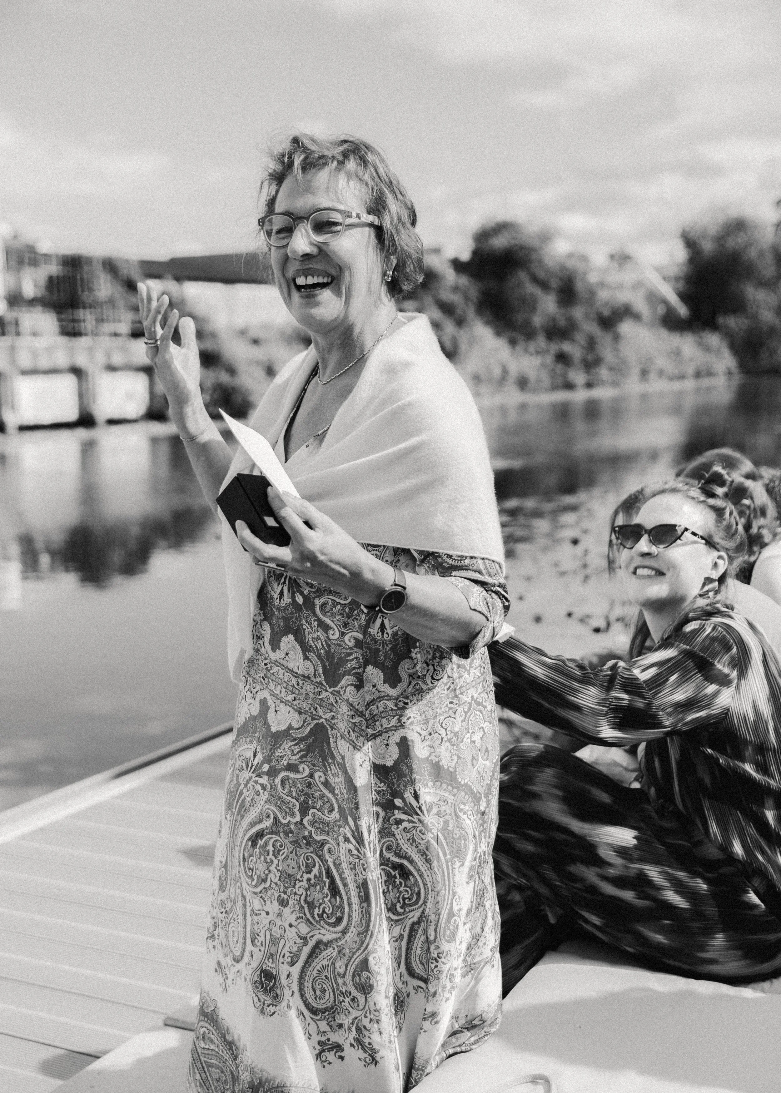 Zwei lachende Frauen sitzen an einem Steg am Fluss, während die ältere Frau etwas liest oder vorträgt, im Hintergrund sind Bäume und ein Himmel mit Wolken zu sehen.