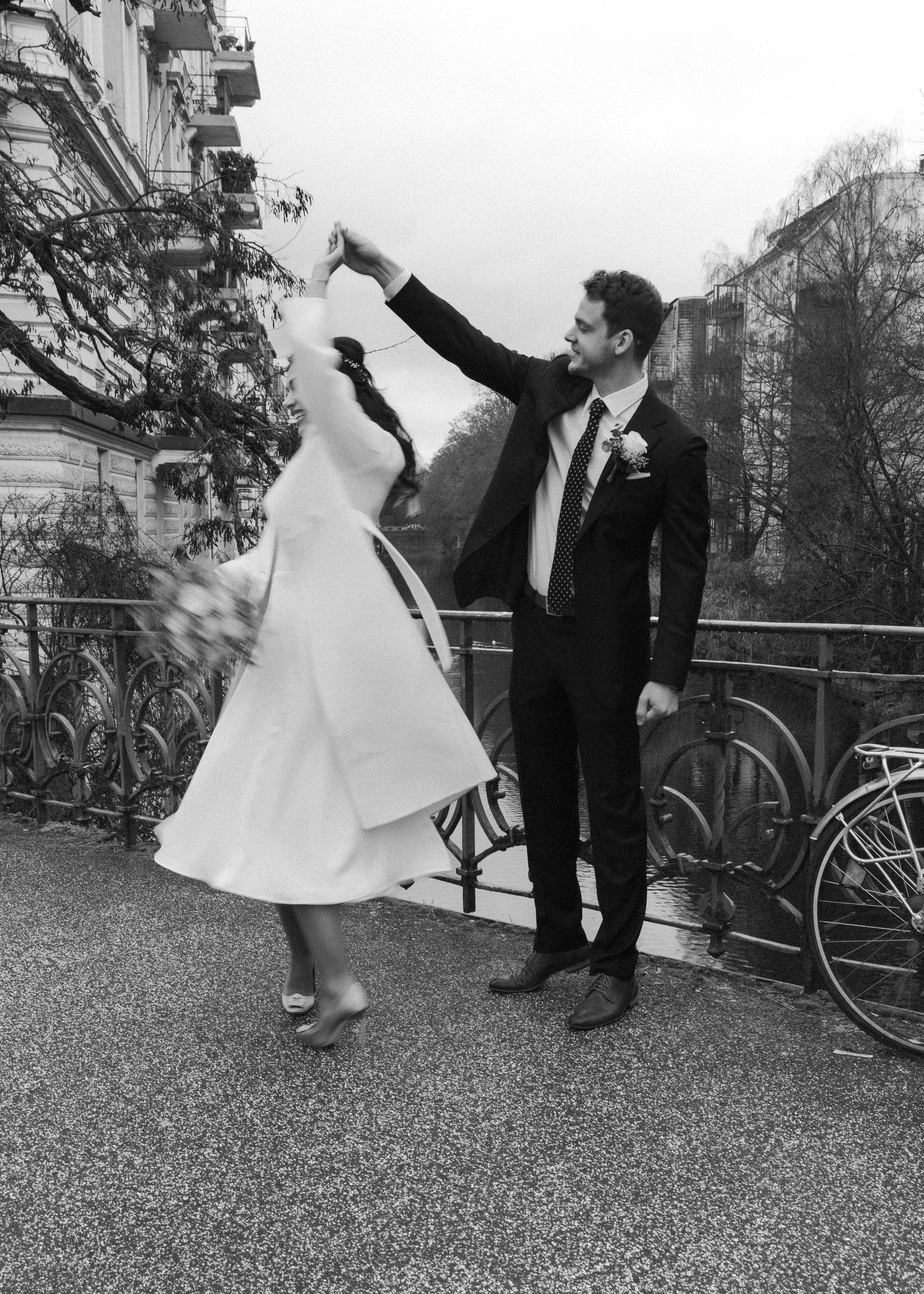 Ein Brautpaar tanzt bei einer Hochzeit im Freien, schwarz-weiß Foto, auf einer Brücke neben einem Fluss, im Hintergrund Bäume und Gebäude, Frau trägt ein Kleid mit Volants, Mann trägt einen Anzug mit Krawatte.