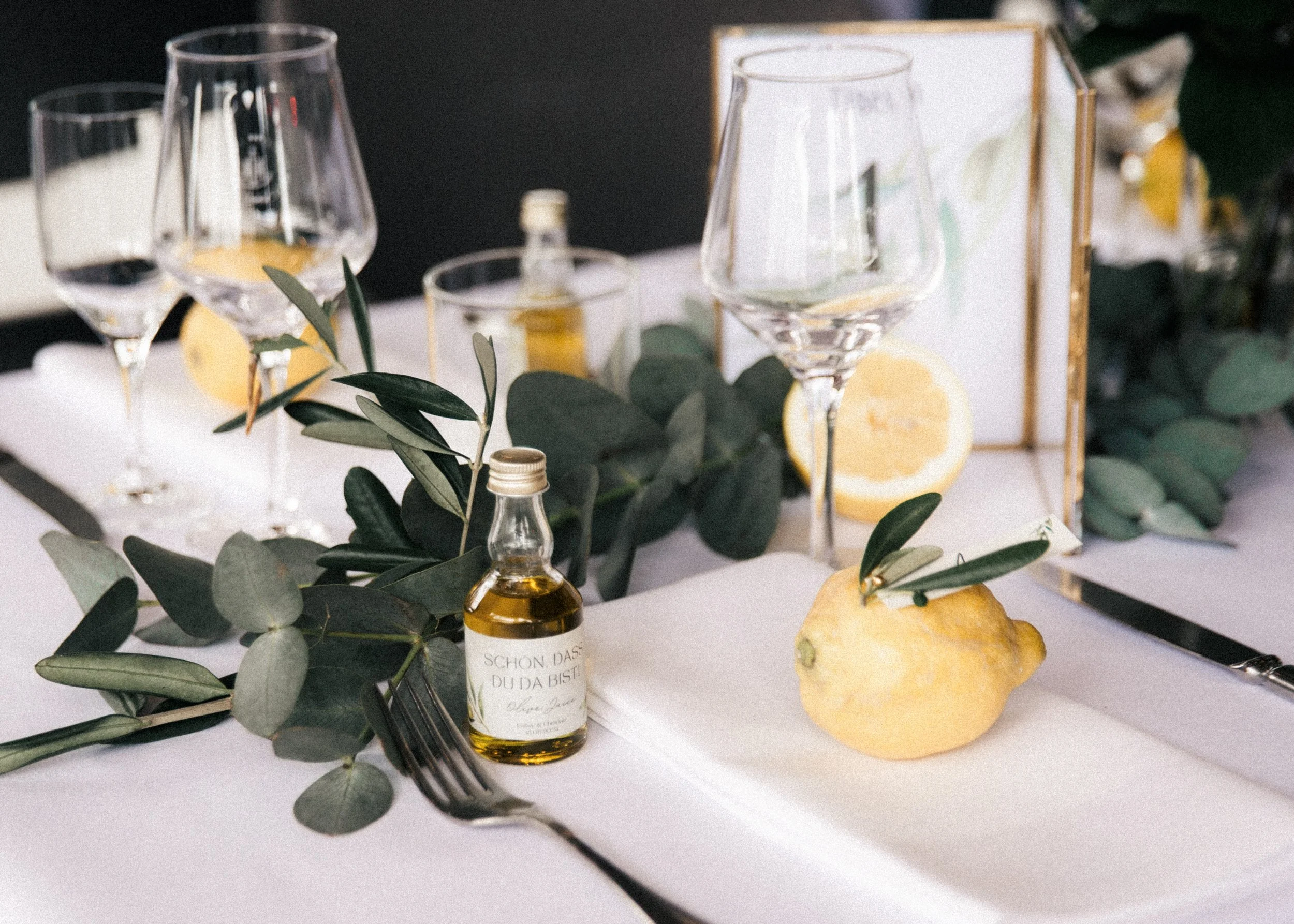 Tischdekoration mit drei Weingläsern, Zitronen, einem kleinen Fläschchen mit Öl, grünen Zweigen, einer weißen Serviette, einem Messer und einer Gabel, auf einer weißen Tischdecke, mit einer dekorativen Box im Hintergrund.