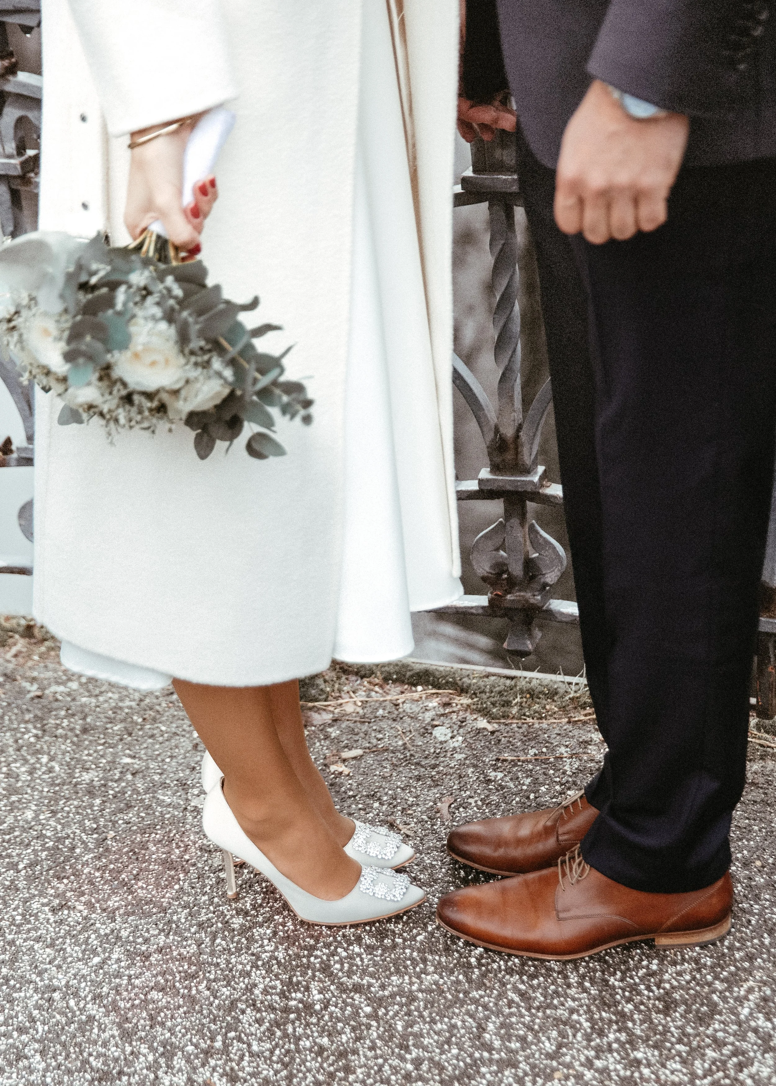 Nahaufnahme der unteren Körperteile eines Paares bei einer formellen Veranstaltung. Die Frau trägt weiße High Heels und einen weißen Rock, während der Mann braune Lederschuhe und dunkle Hosen trägt. Die Begleitung hält einen Blumenstrauß und steht au