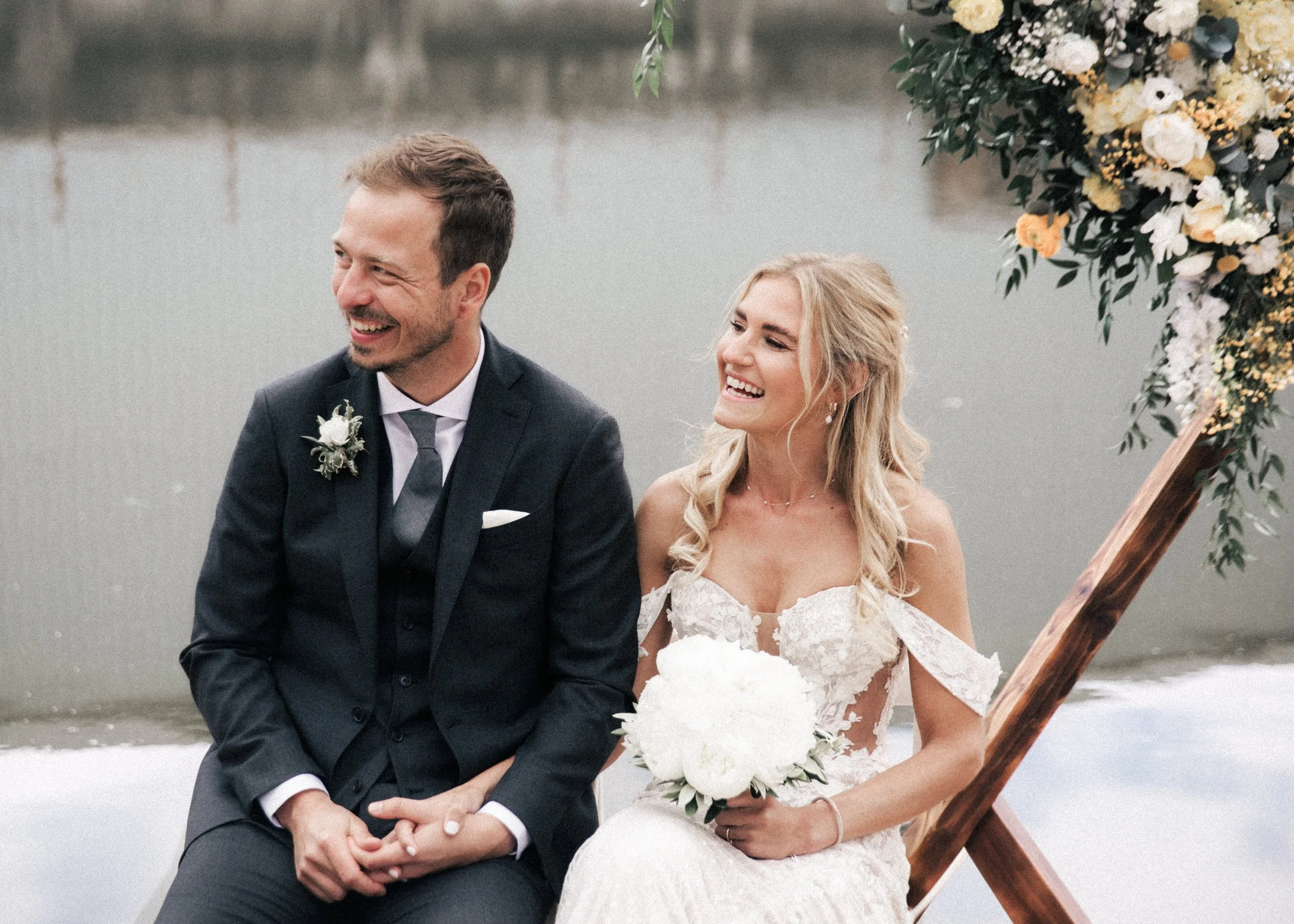 Ein glückliches Brautpaar bei ihrer Hochzeitszeremonie am Wasser, der Bräutigam in einem dunklen Anzug und die Braut in einem weißen, trägerlosen Kleid, beide lachen und halten sich an den Händen.