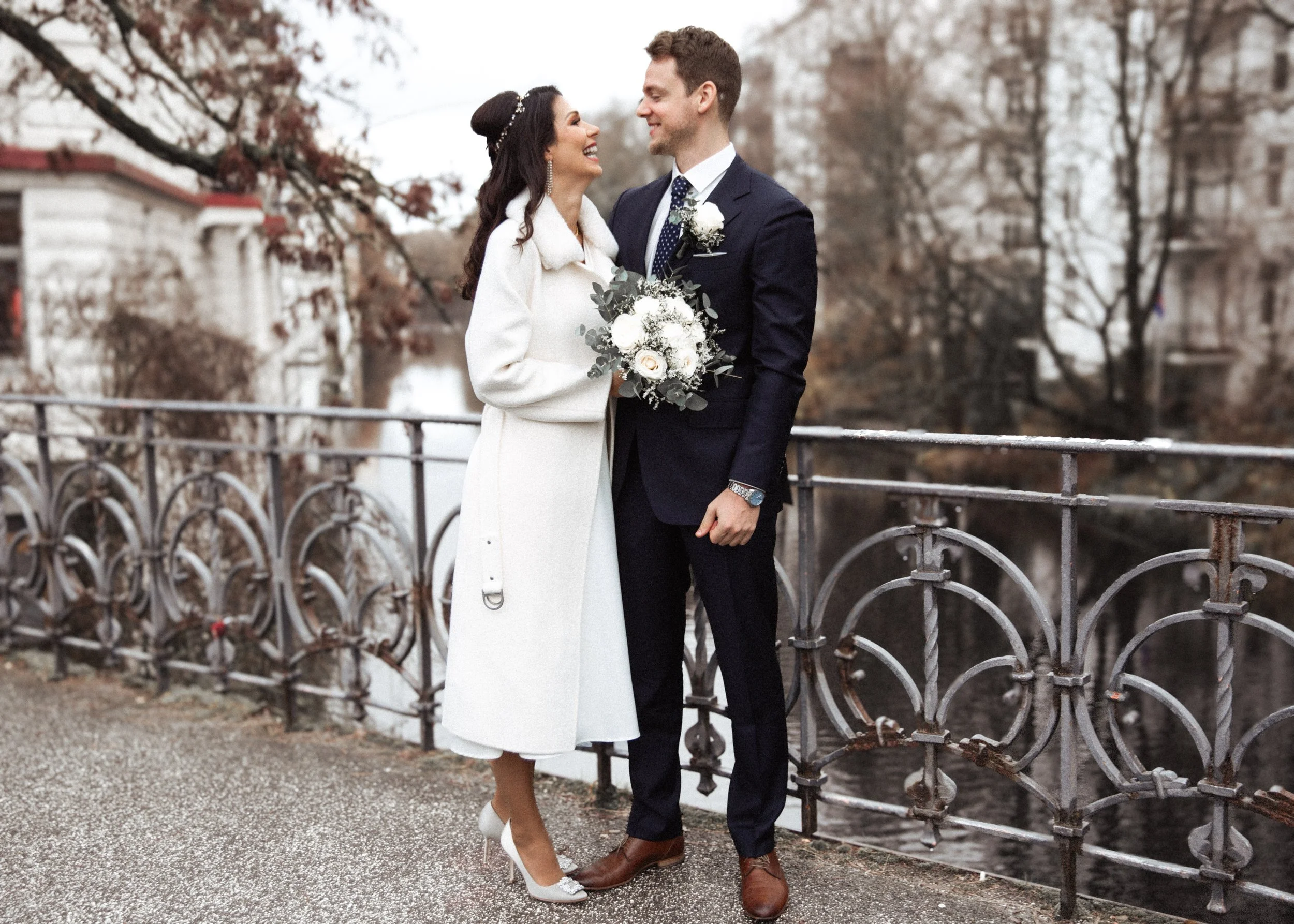 Ein Brautpaar steht am Flussufer vor einer Metallbrüstung, lächelnd und sich süß zuwinkend, bei ihrer Hochzeit im Herbst.