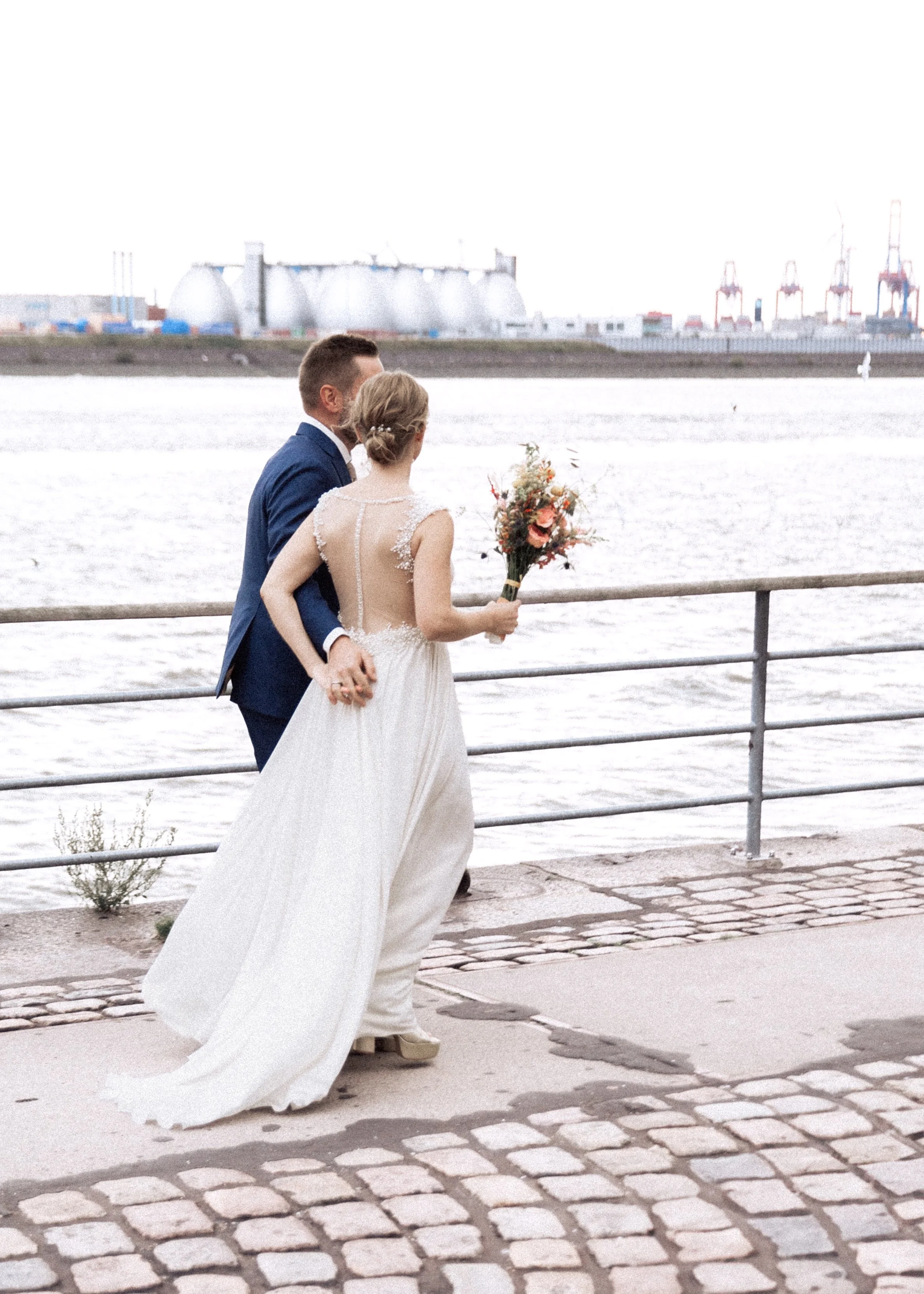 Ein Brautpaar an einem Flussufer während einer Hochzeit, die Braut hält einen Blumenstrauß, im Hintergrund Industrieanlagen und Wasser.