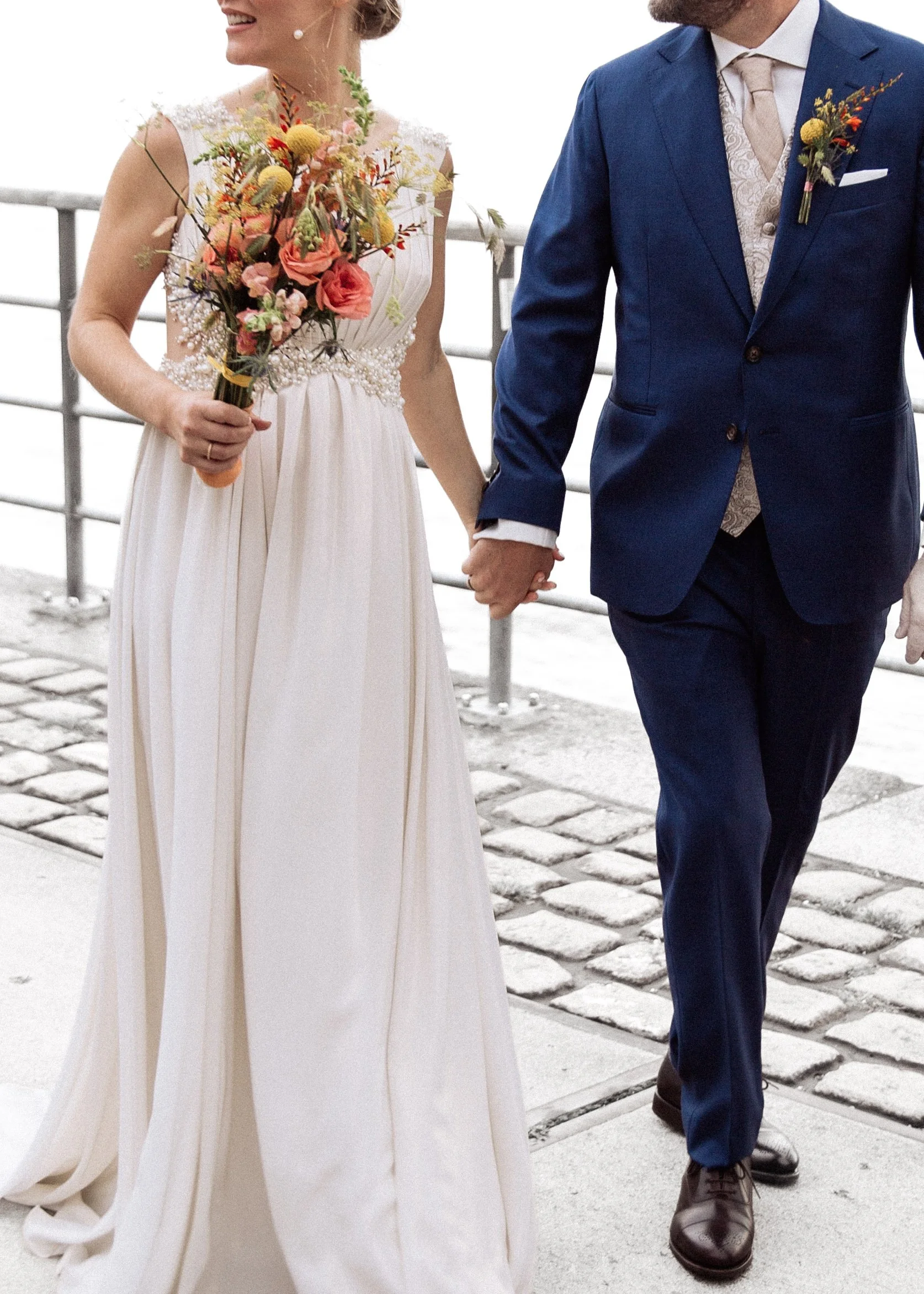 Ein Brautpaar hält sich an den Händen bei einer Hochzeit am Wasser, die Braut trägt ein langes weißes Kleid und hält einen bunten Blumenstrauß, der Bräutigam trägt einen blauen Anzug mit beige Weste und Krawatte.