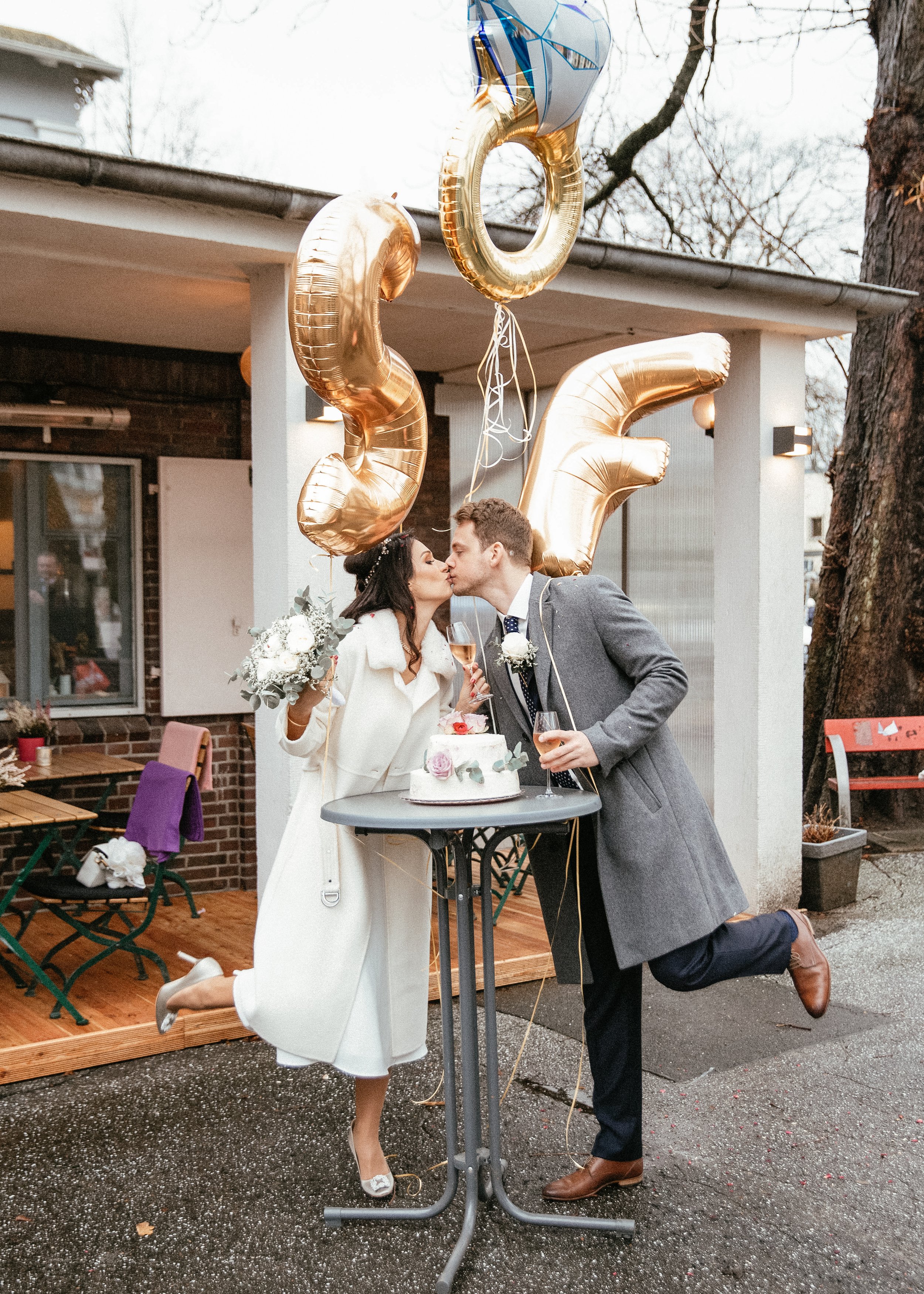 Ein Brautpaar küsst sich vor einem Hochzeitstisch mit einer Torte bei einer Feier im Freien, umgeben von goldenen Ballons, die die Zahl 30 bilden.