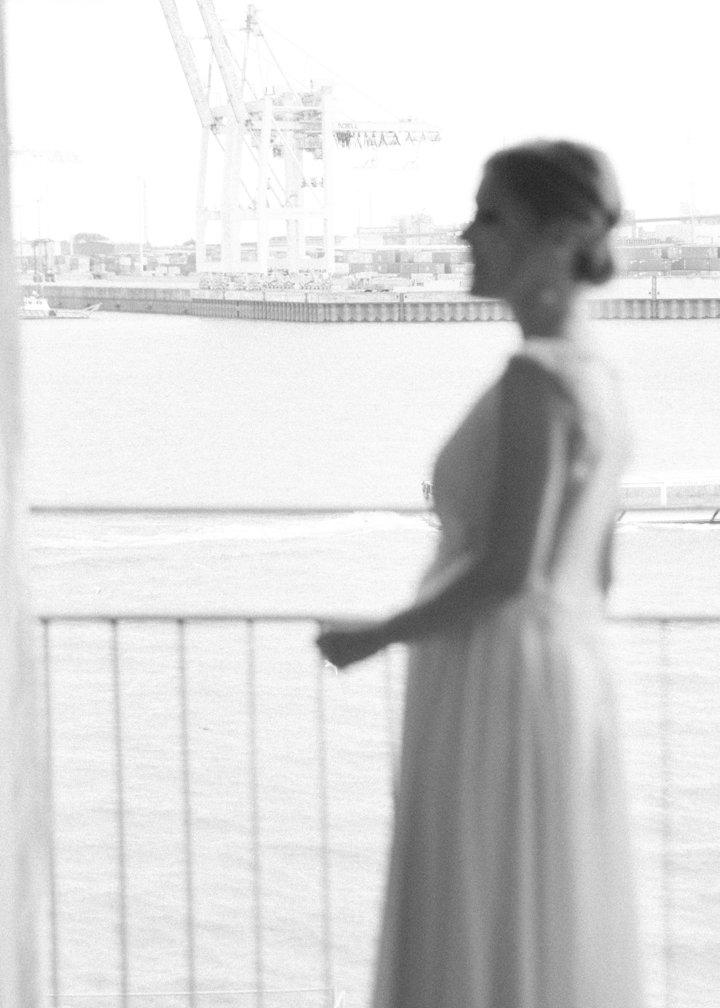 Eine Frau in einem Kleid steht auf einem Balkon am Wasser mit einer Hafenlandschaft im Hintergrund.