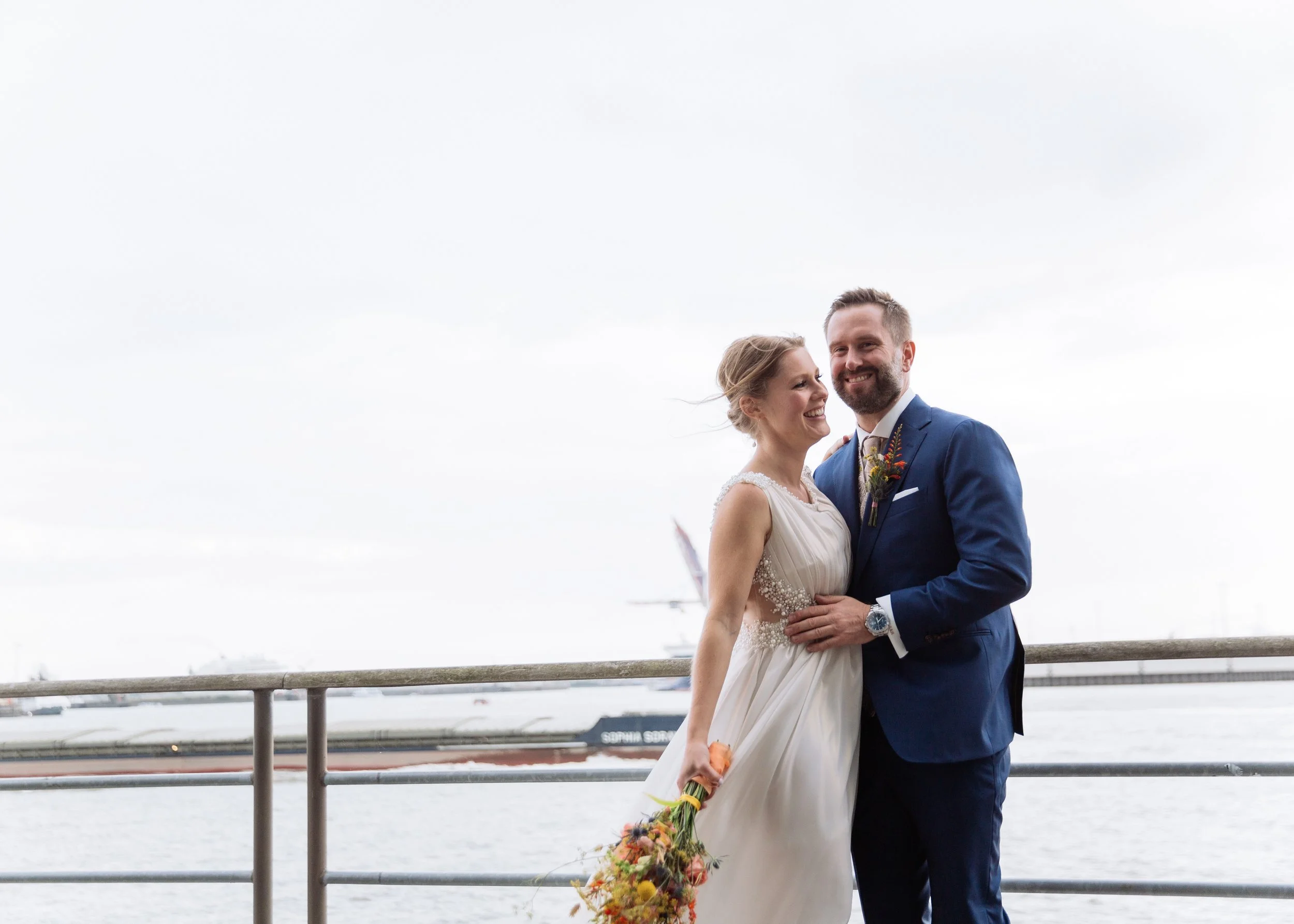 Ein lachendes Brautpaar steht auf einem Bootsterminal mit Wasser und Schiffen im Hintergrund, die Braut hält einen Blumenstrauß.
