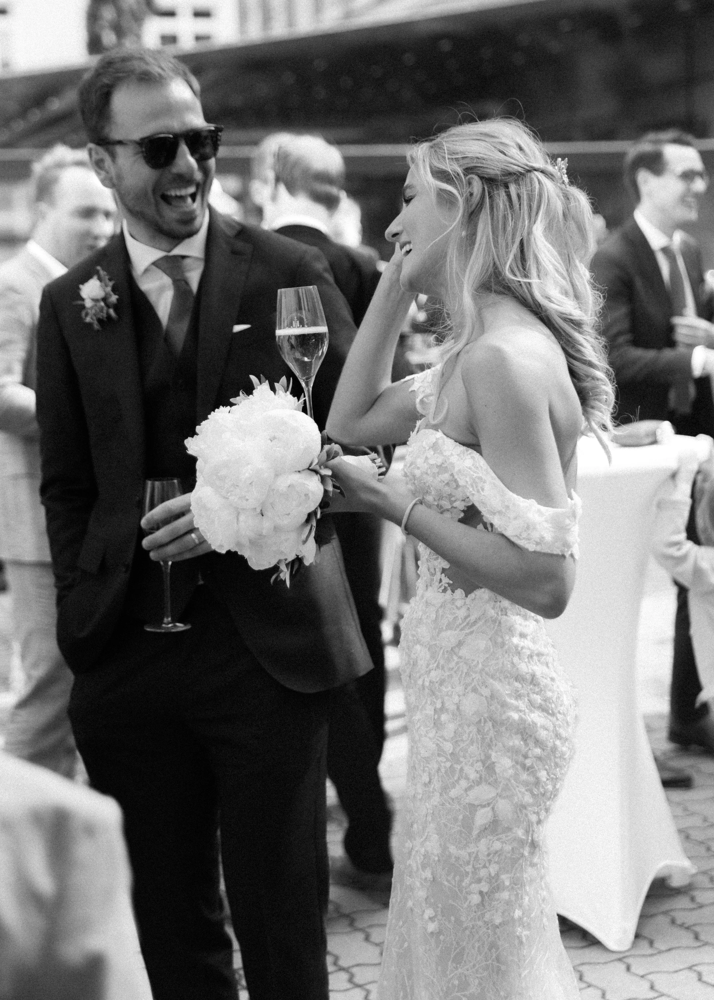 Braut und Bräutigam bei ihrer Hochzeitsfeier, der Bräutigam hält ein Glas Champagner und ein Blumenstrauß, beide lachen und genießen den Moment.