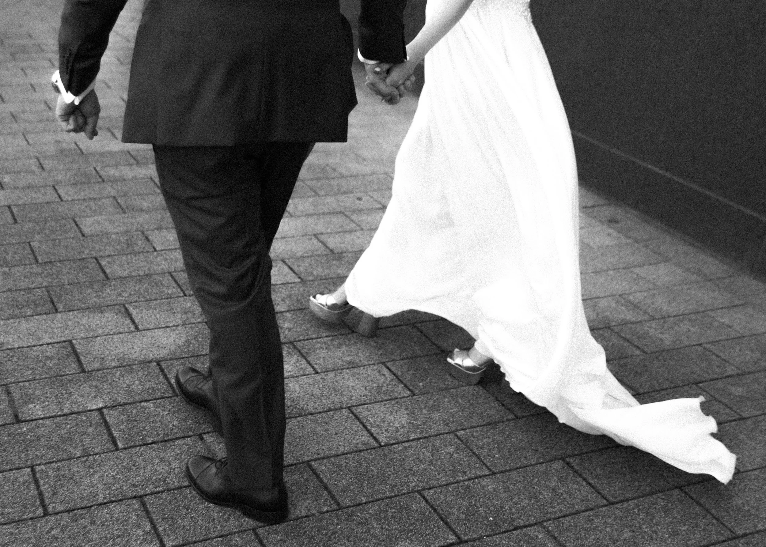 Nahaufnahme eines Paares, das sich bei ihrer Hochzeit anfasst, mit dem Fokus auf ihre Füße und Hände, die auf gepflastertem Boden stehen.