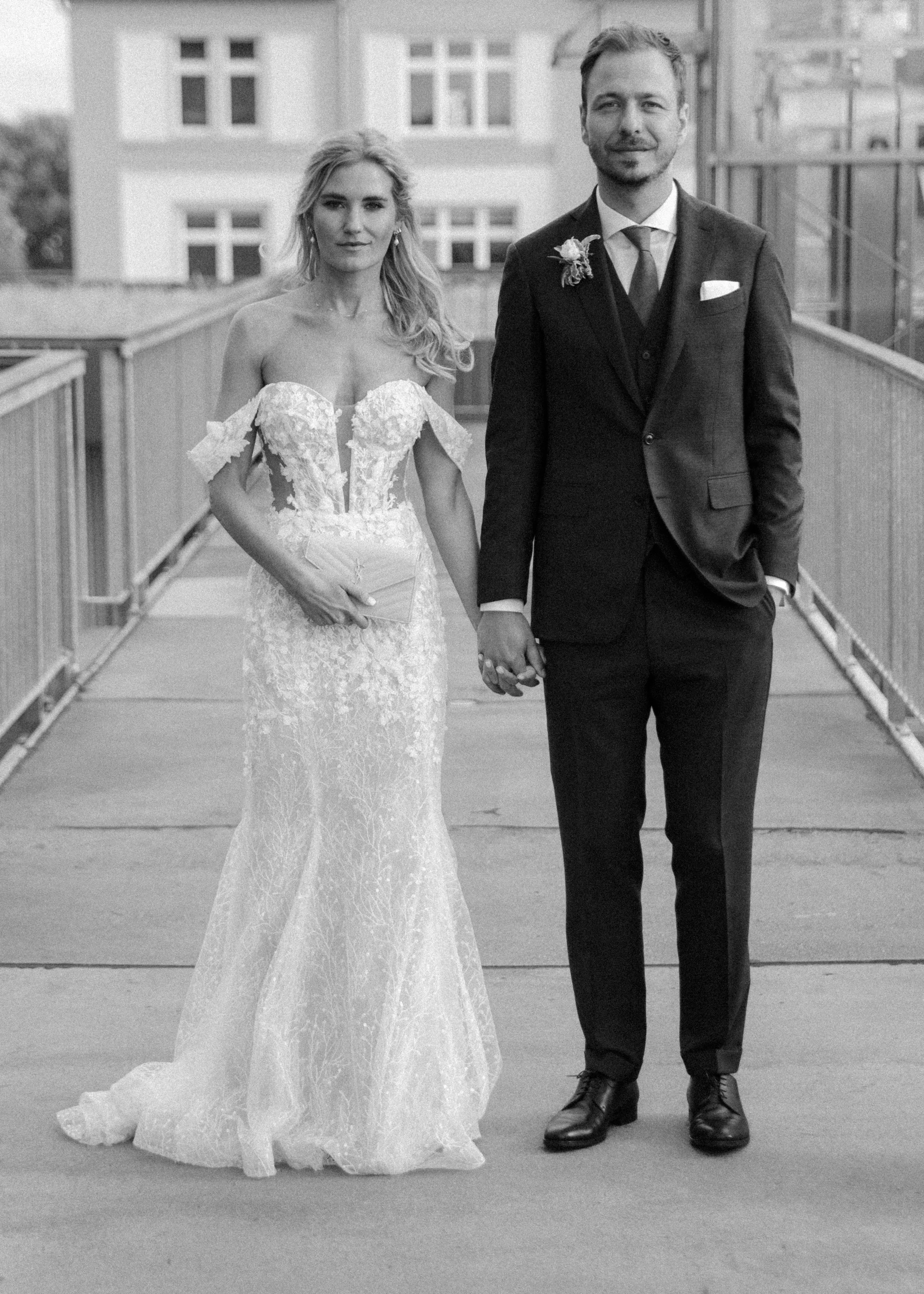Ein Brautpaar in Hochzeitskleidung, das einander auf einer Brücke hält, im Hintergrund ein mehrstöckiges Gebäude, schwarz-weiß Foto.