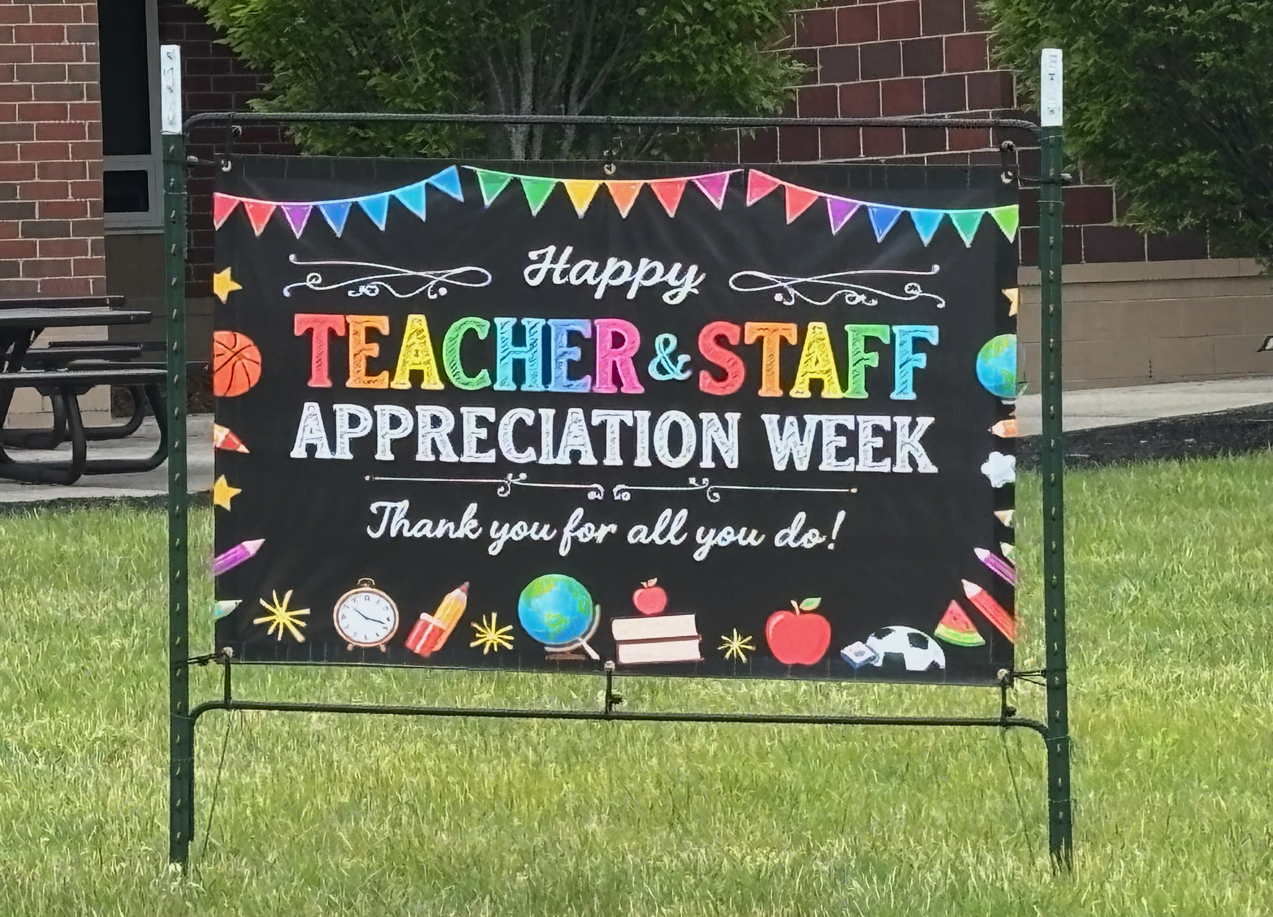 A black banner with colorful text and drawings celebrating Teacher & Staff Appreciation Week, decorated with paper bunting, stars, globes, a clock, apples, and other school-themed illustrations.