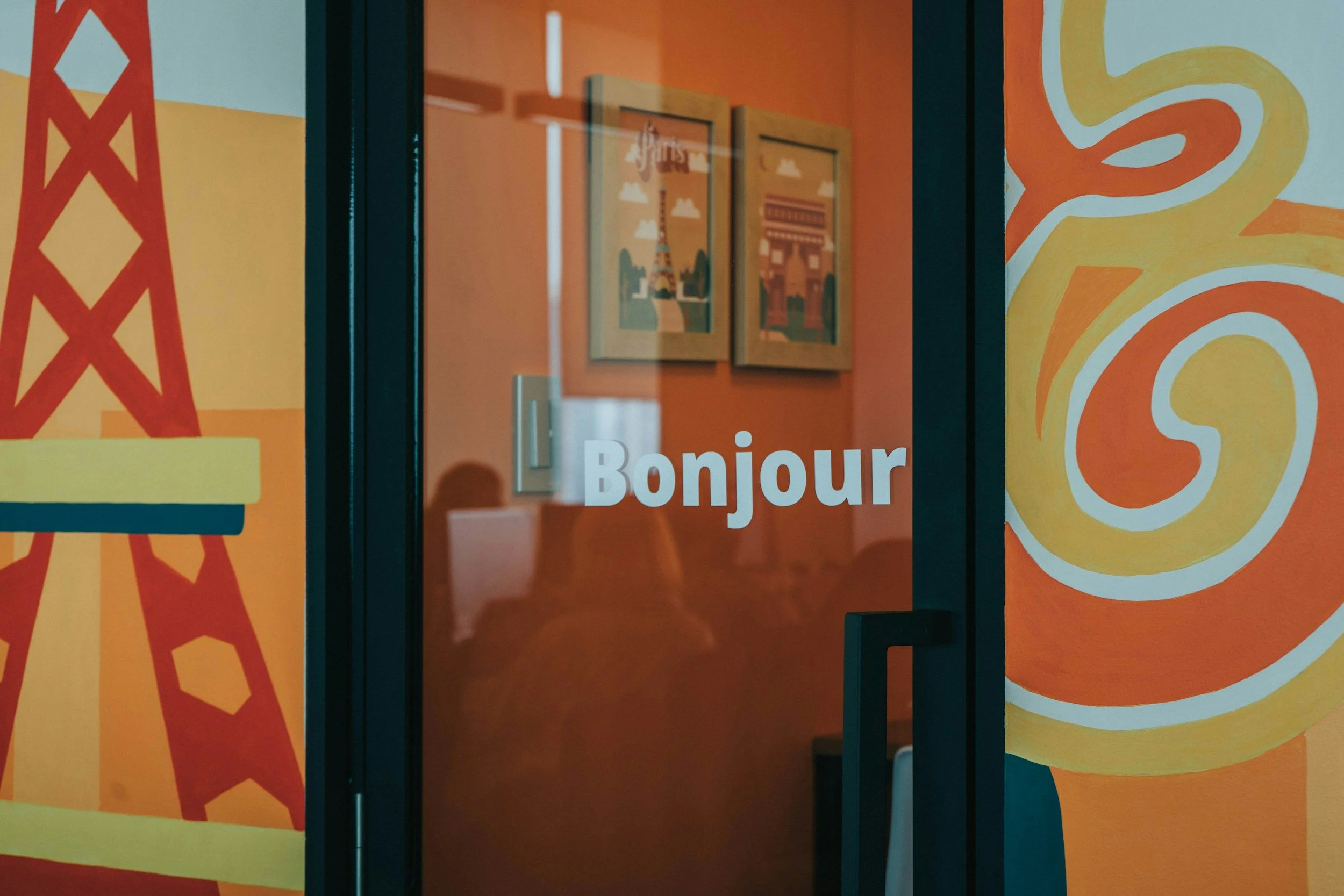Colorful mural on wall with drawings of the Eiffel Tower and Arc de Triomphe, and the word 'Bonjour' on a glass door.